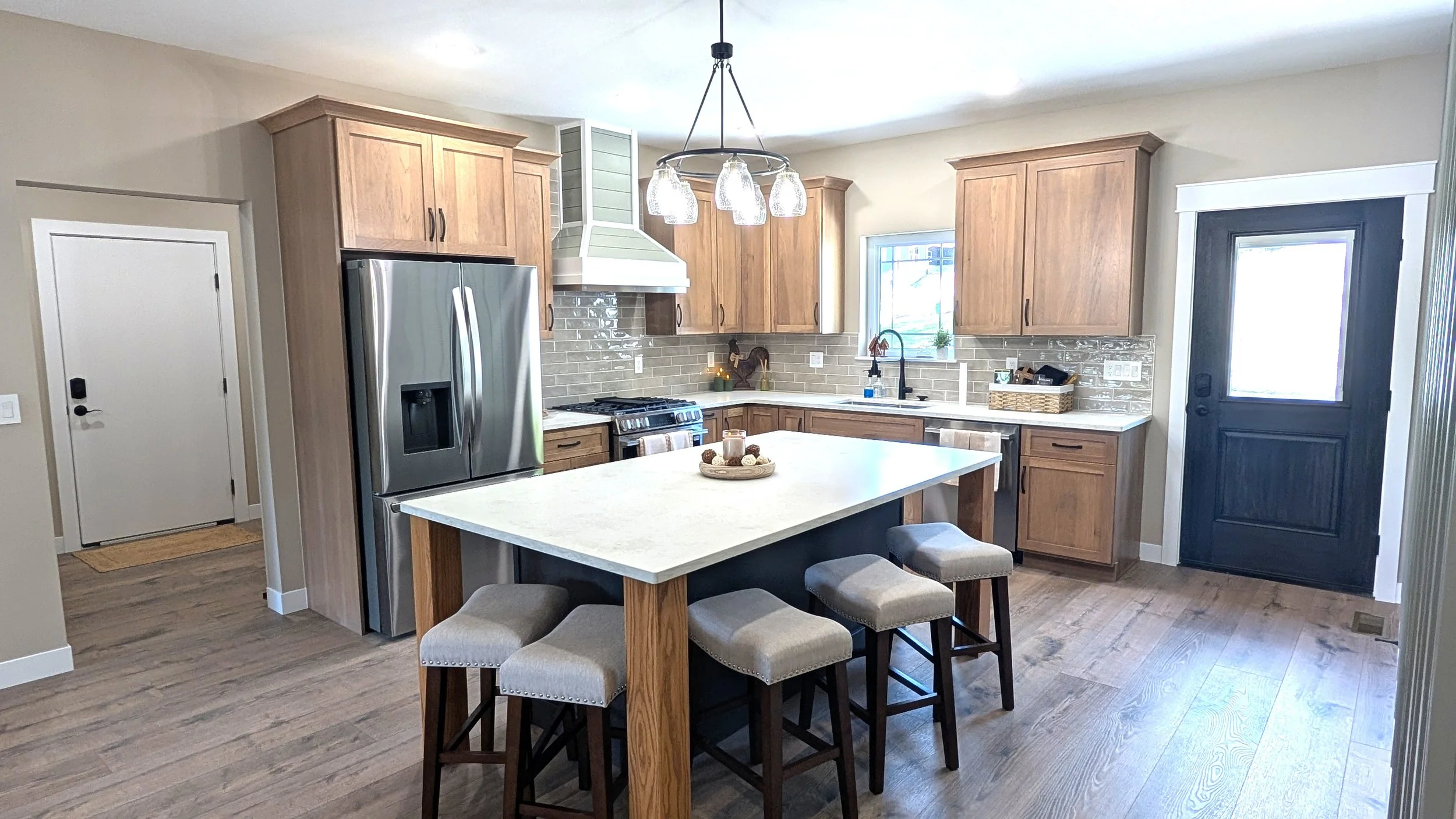 A modern kitchen with wooden cabinets, stainless steel refrigerator, black stove, and a central island with beige stools. There is a chandelier overhead, a window above the sink, and a door to the right. The kitchen has light-colored walls and wood flooring.