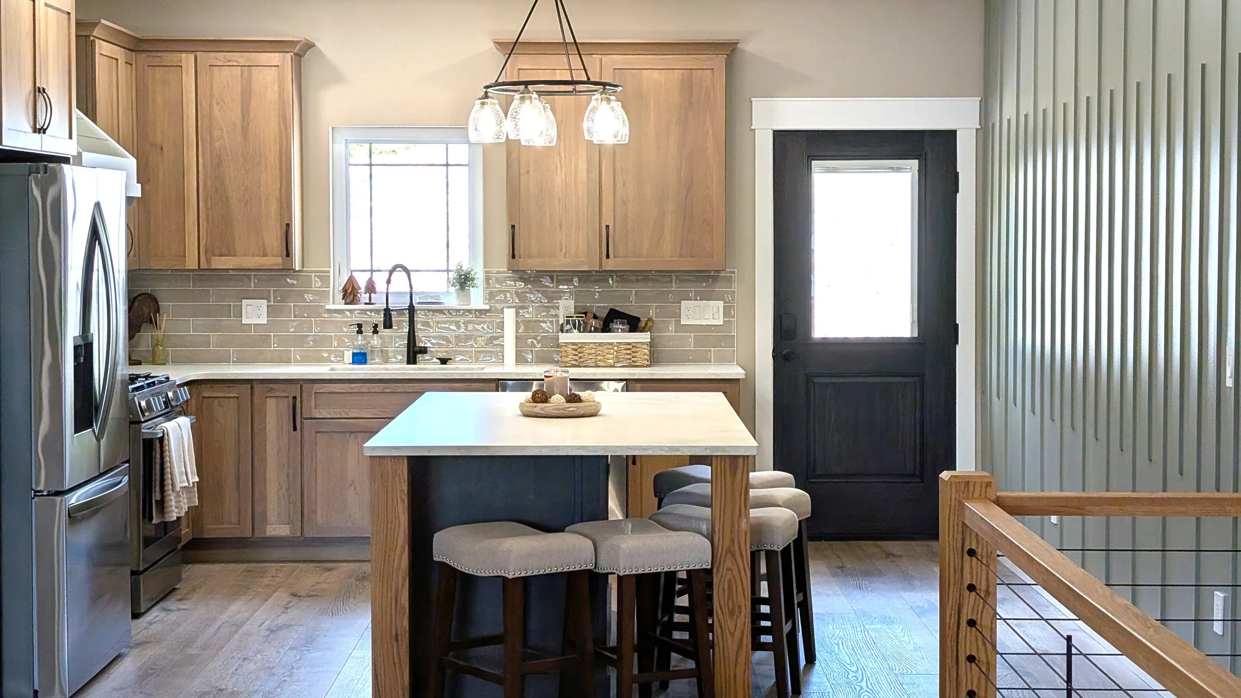 A modern kitchen with wooden cabinets, a stainless steel refrigerator, a stove, and a window above the sink. There is an island with barstools, a round chandelier, and a dark door leading outside. The wall on the right has vertical paneling.