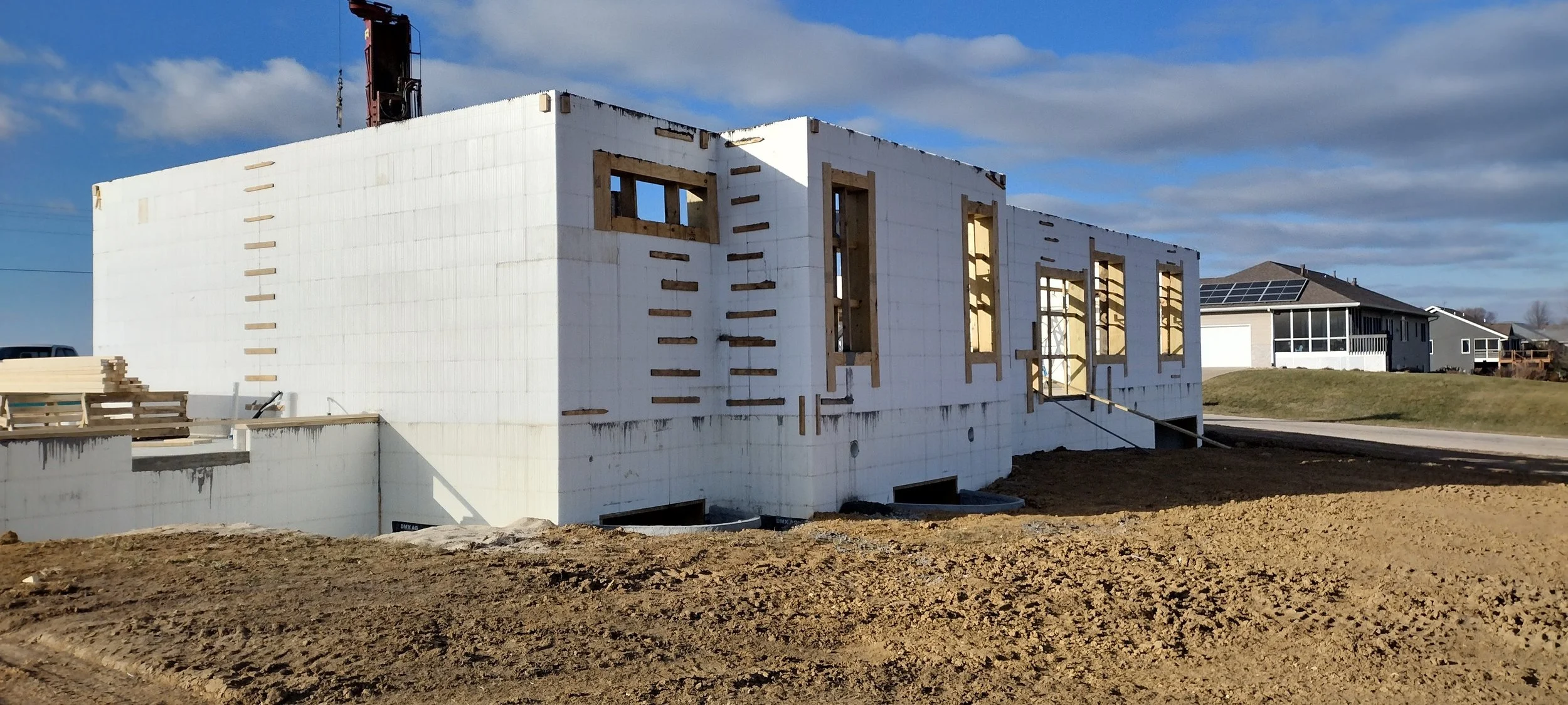 A house under construction with insulated concrete form (ICF) walls and window openings, set against a blue sky with clouds, neighboring houses visible in the background, and construction materials and dirt in the foreground.