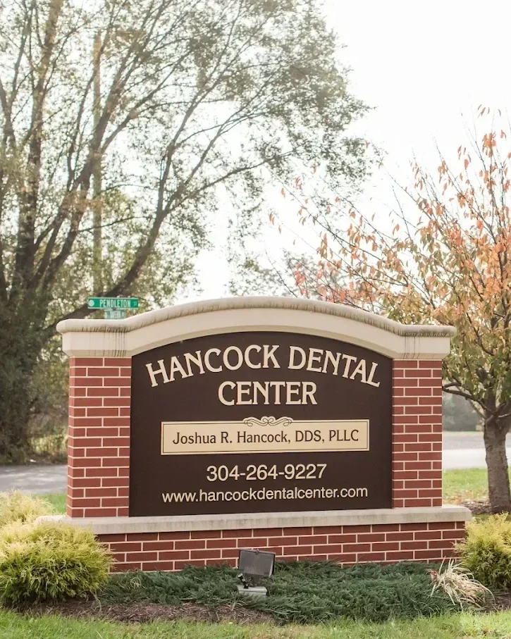 Sign for Hancock Dental Center with contact information and website, surrounded by landscaped bushes, trees in the background, and a street sign at the corner.