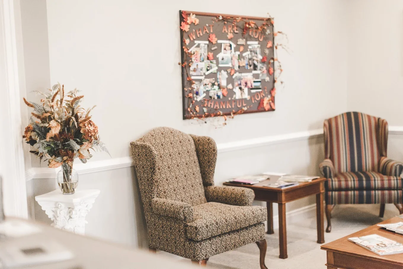 Waiting room with two patterned armchairs, a side table with magazines, a coat rack with magazines on top, and a decorated wall display with pictures and a sign that reads "What are you thankful for?"