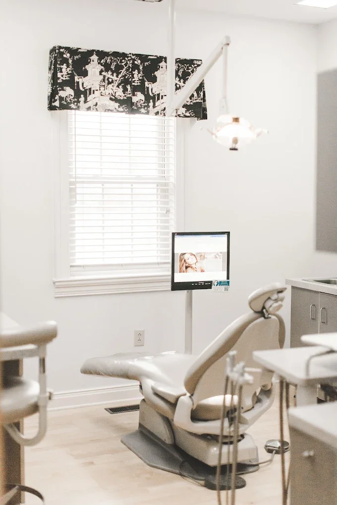 Empty dental examination room with a dental chair, monitor, and dental tools, window with black and white patterned valance, and a small cabinet.