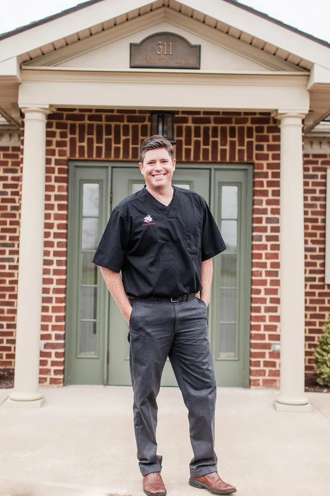 Dr. Hancock in medical scrubs standing in front of a brick house with green double doors and white columns.