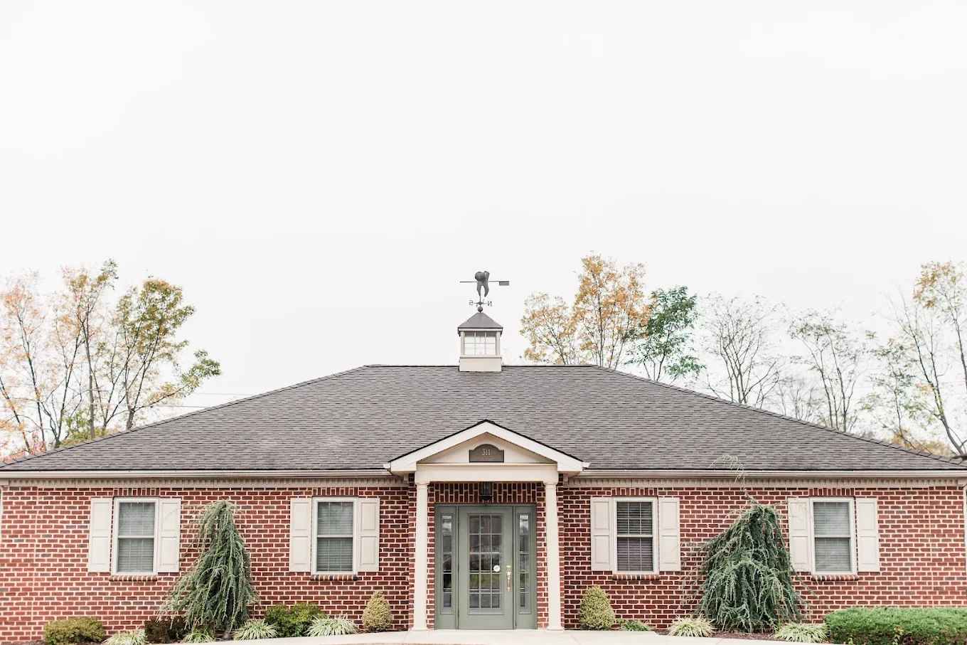 A brick building with a gray door, white shutters, and small bushes in front, with trees in the background and a weather vein on the roof.