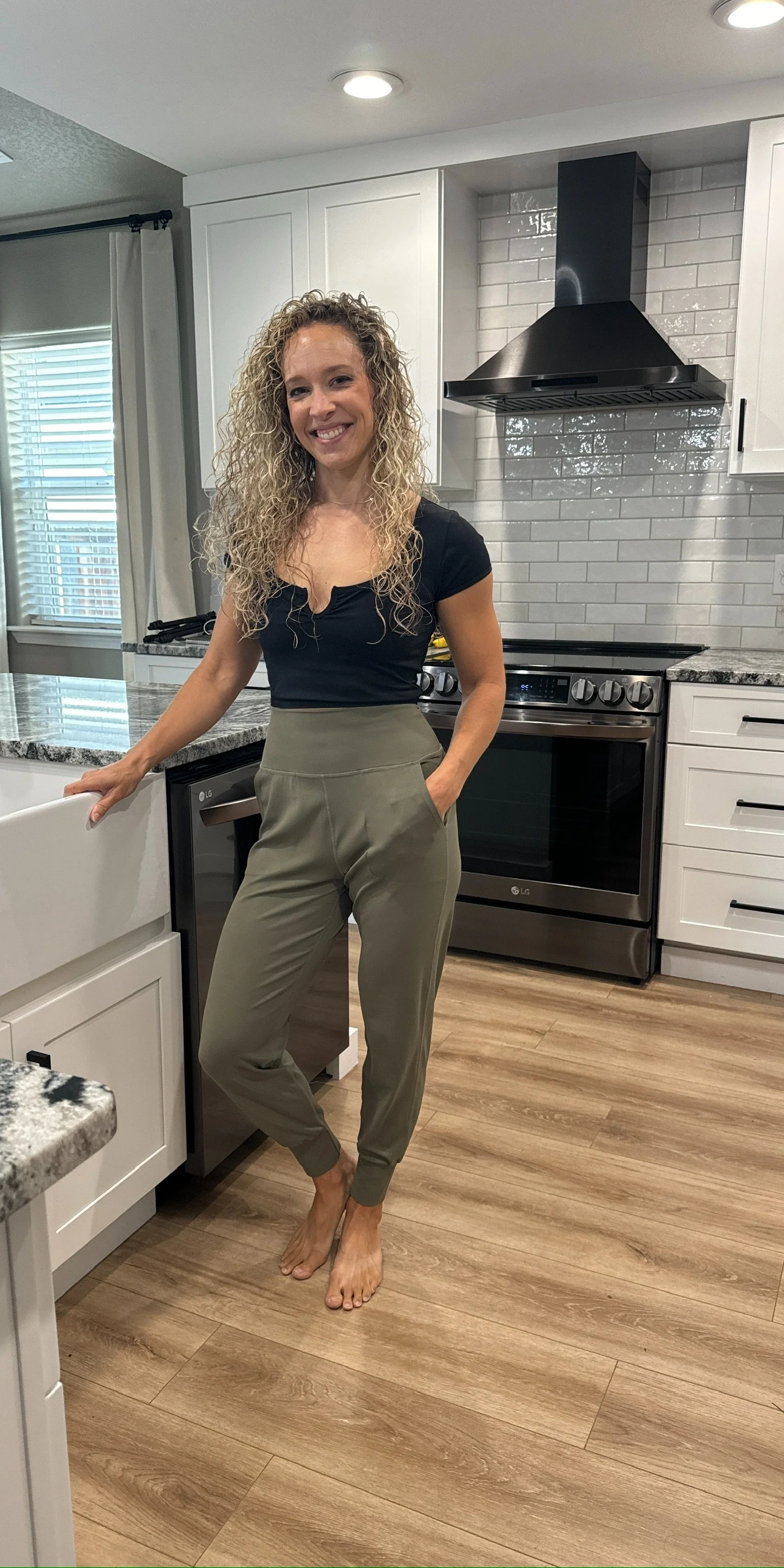 A woman with blonde curly hair smiling in a modern kitchen, wearing a black top and beige pants, barefoot, standing next to a kitchen counter and a stove.