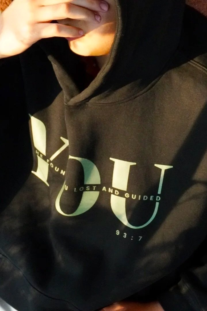 Person wearing a black hoodie with the words "He’s Found You Lost and Guided YOU" the YOU is in big letters across the shirt and the numbers "93:7" printed on it, taking a selfie with a hand partially covering their face.