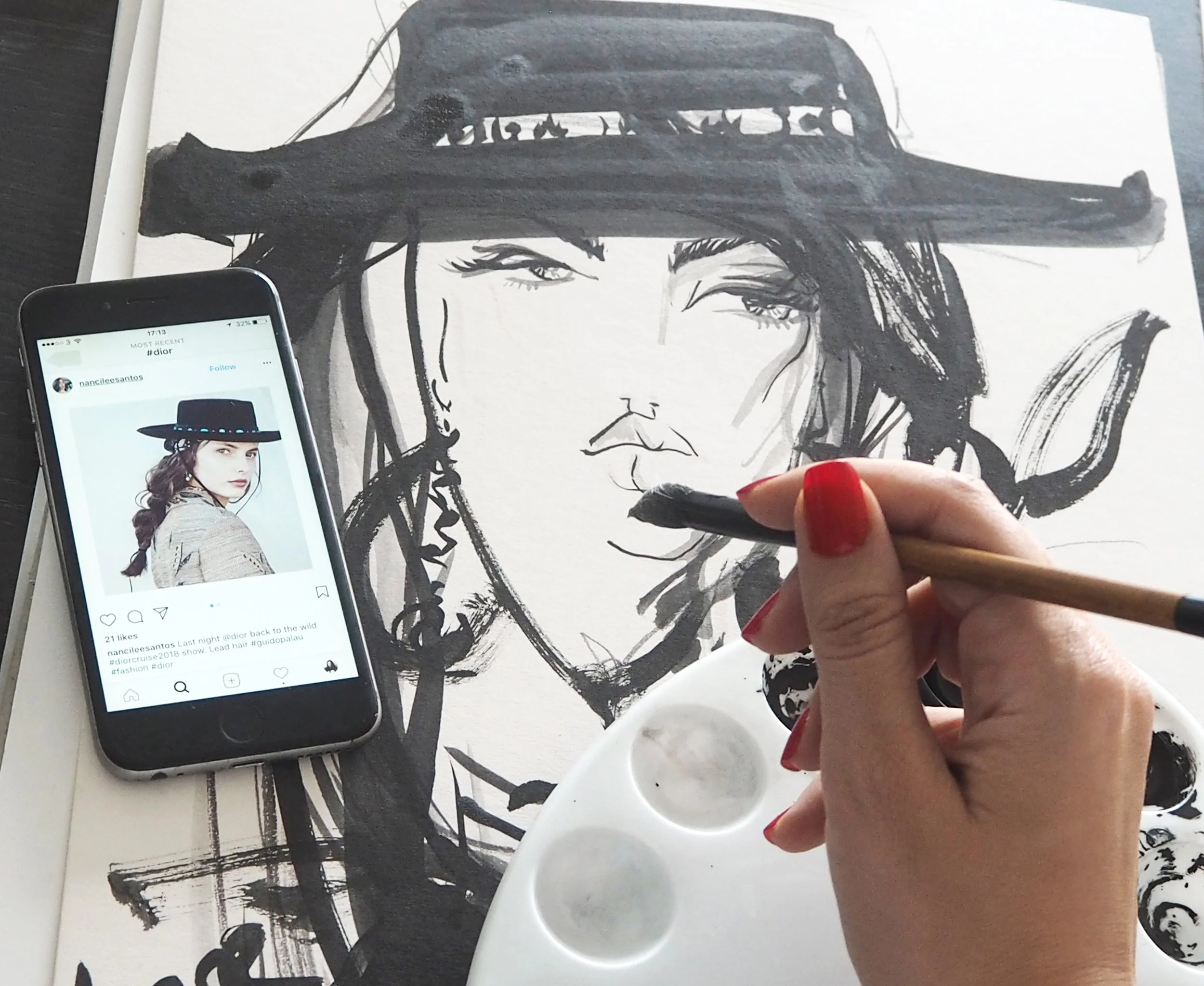 Hand with painted nails holding a paintbrush, painting a black and white fashion illustration of a woman wearing a fedora hat, with a smartphone displaying the same fashion illustration and Instagram post.