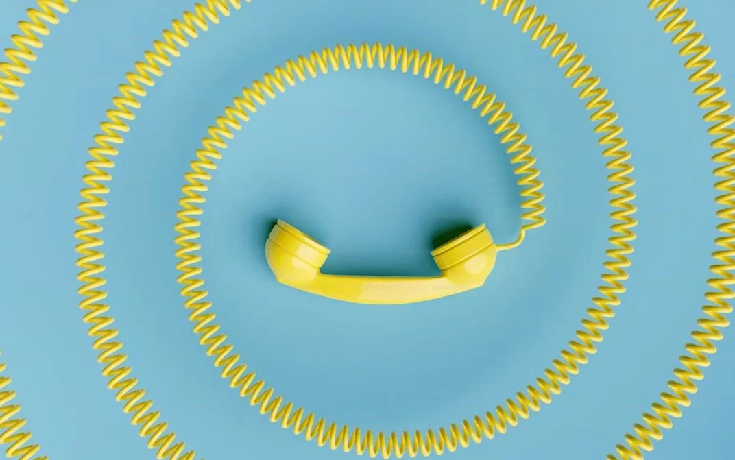 Yellow telephone handset arranged to look like a smiley face, with spiral cords around it on a blue background.