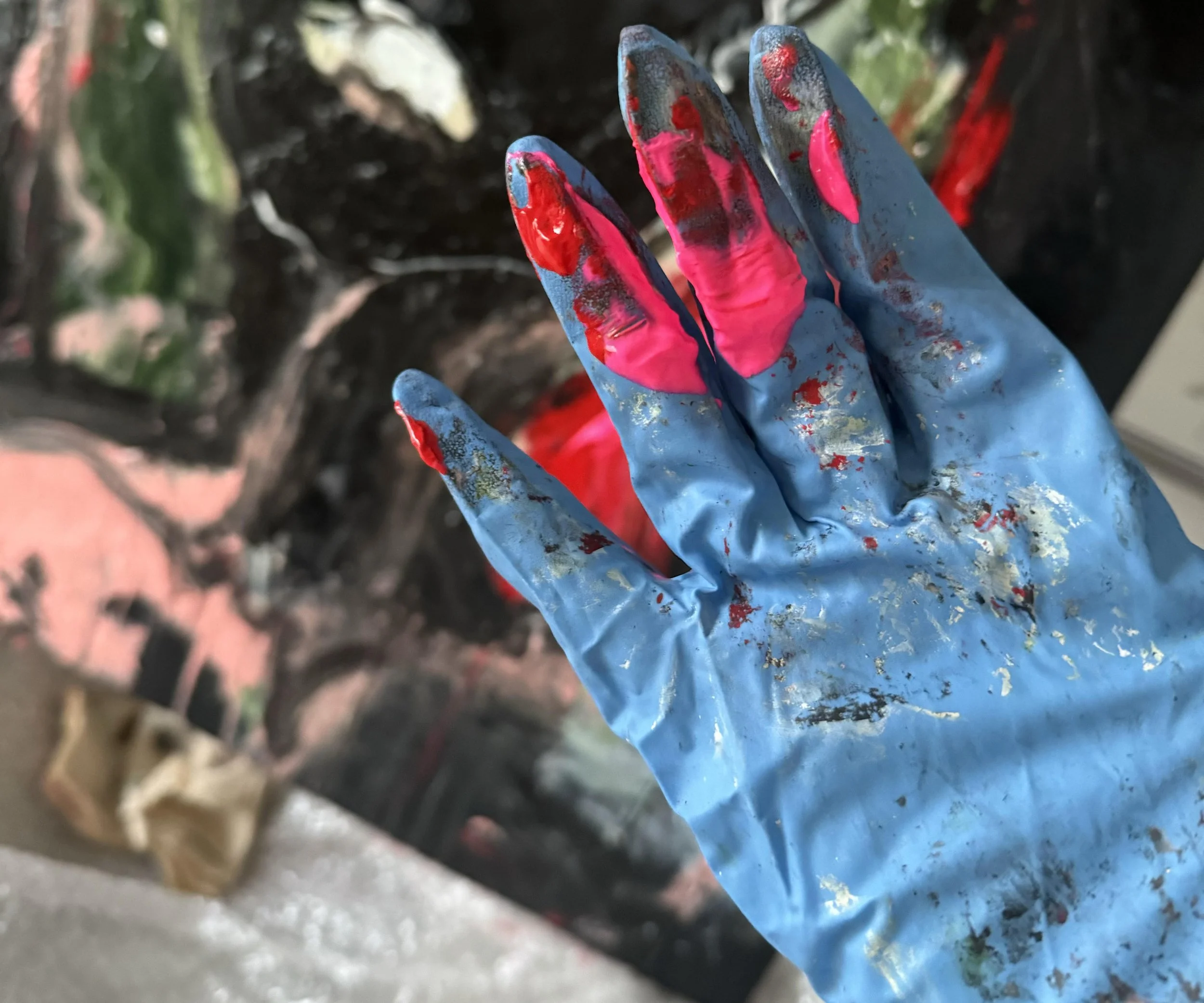 Close-up of a gloved hand covered in wet, dripping paint, with the background showing blurred greenery and soil.