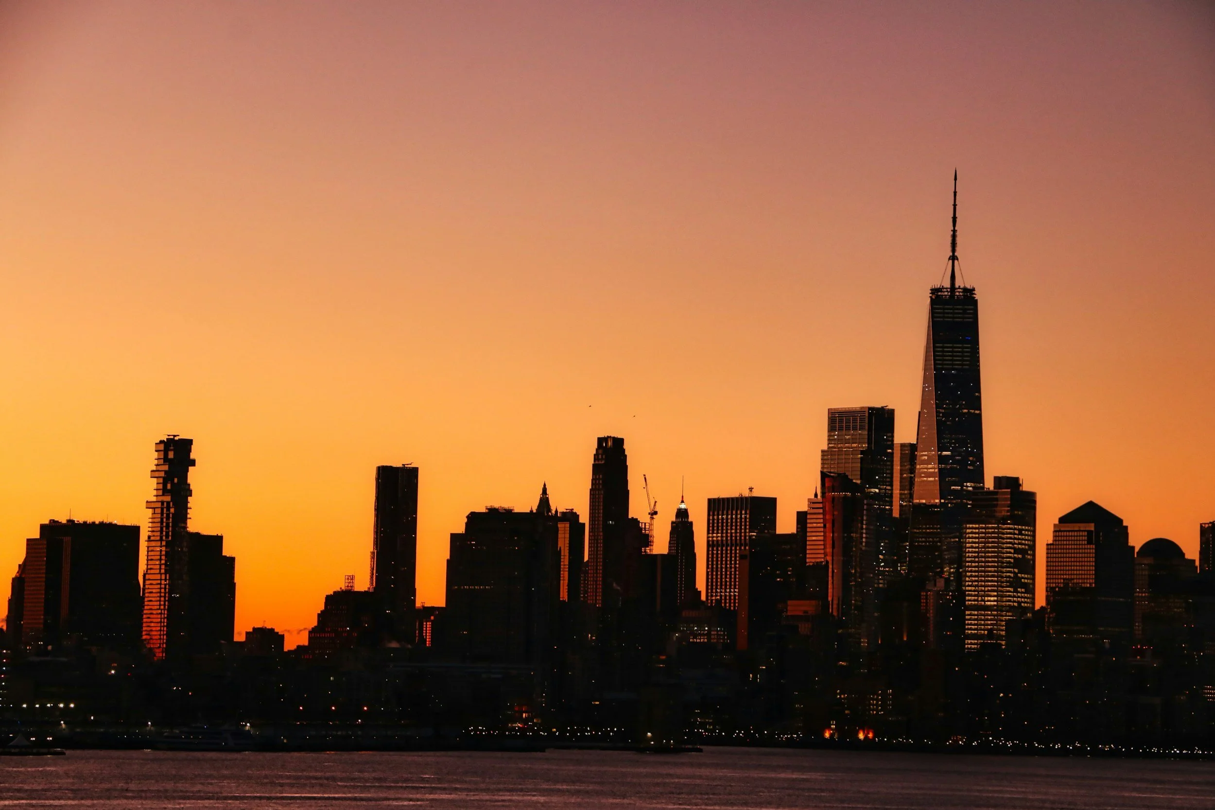 Silhouette of New York City skyline during sunset with tall buildings and One World Trade Center visible.