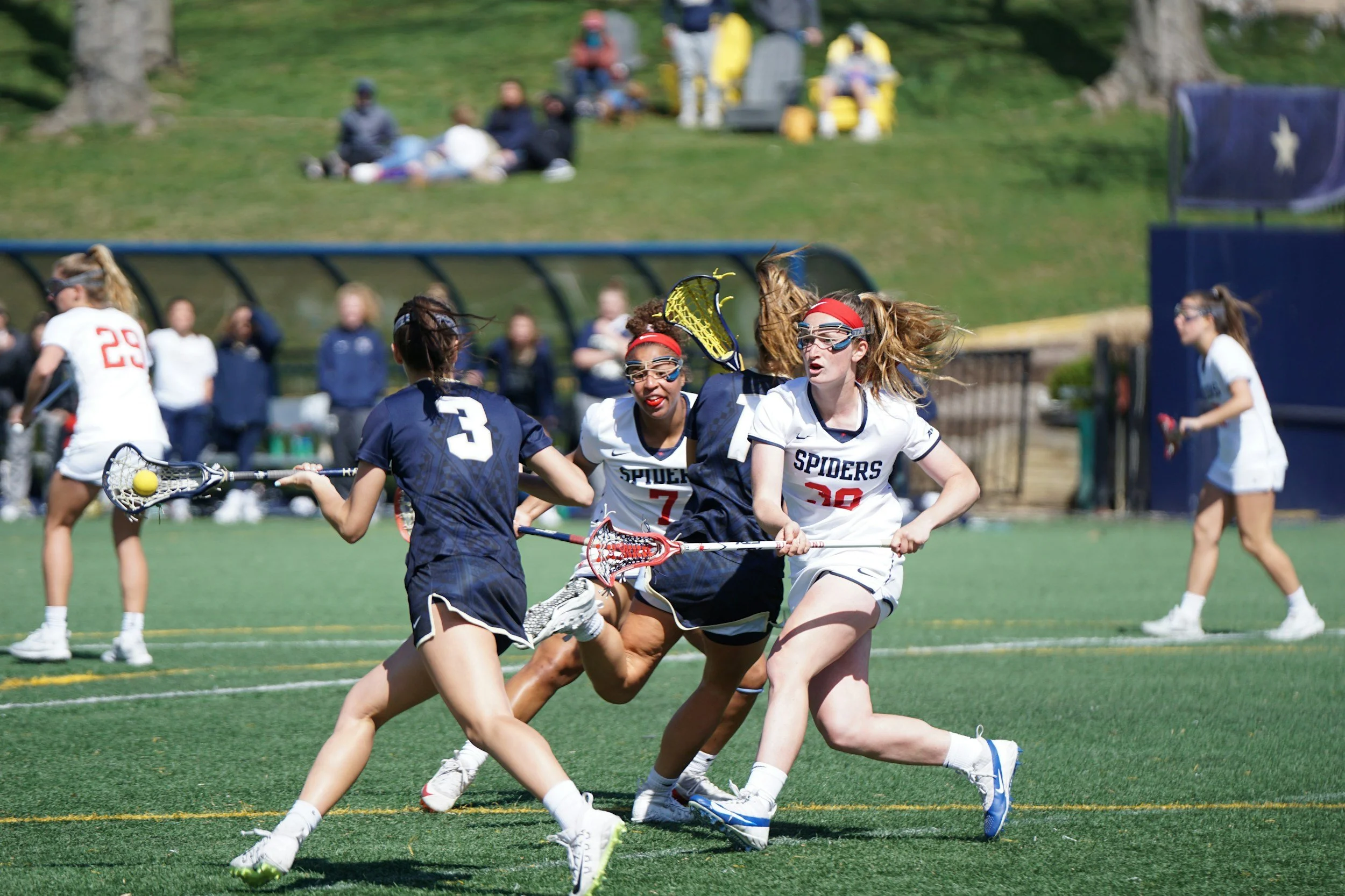 Lacrosse players in action during a game on a green field, with spectators and trees in the background.