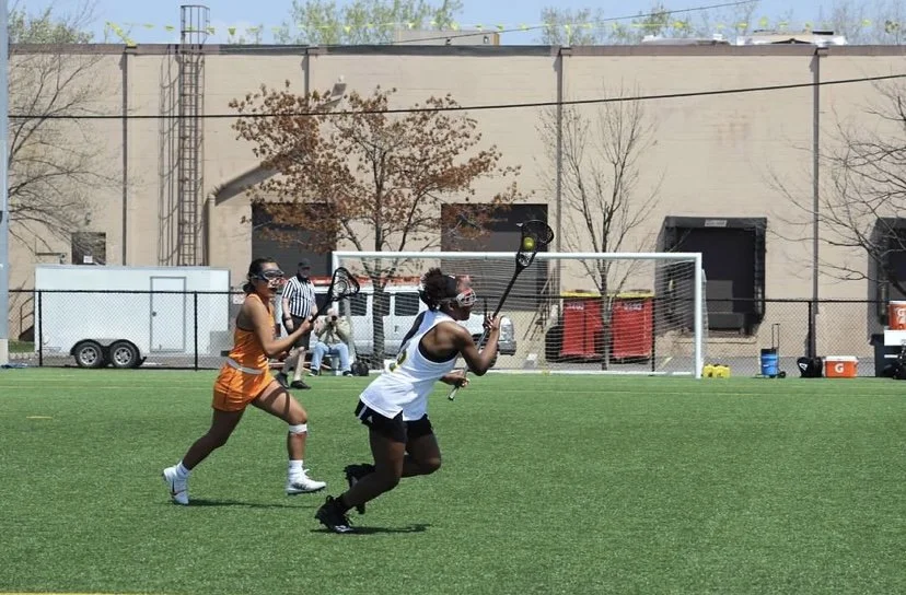 Two women playing lacrosse on a green field, with a soccer goal and building in the background.