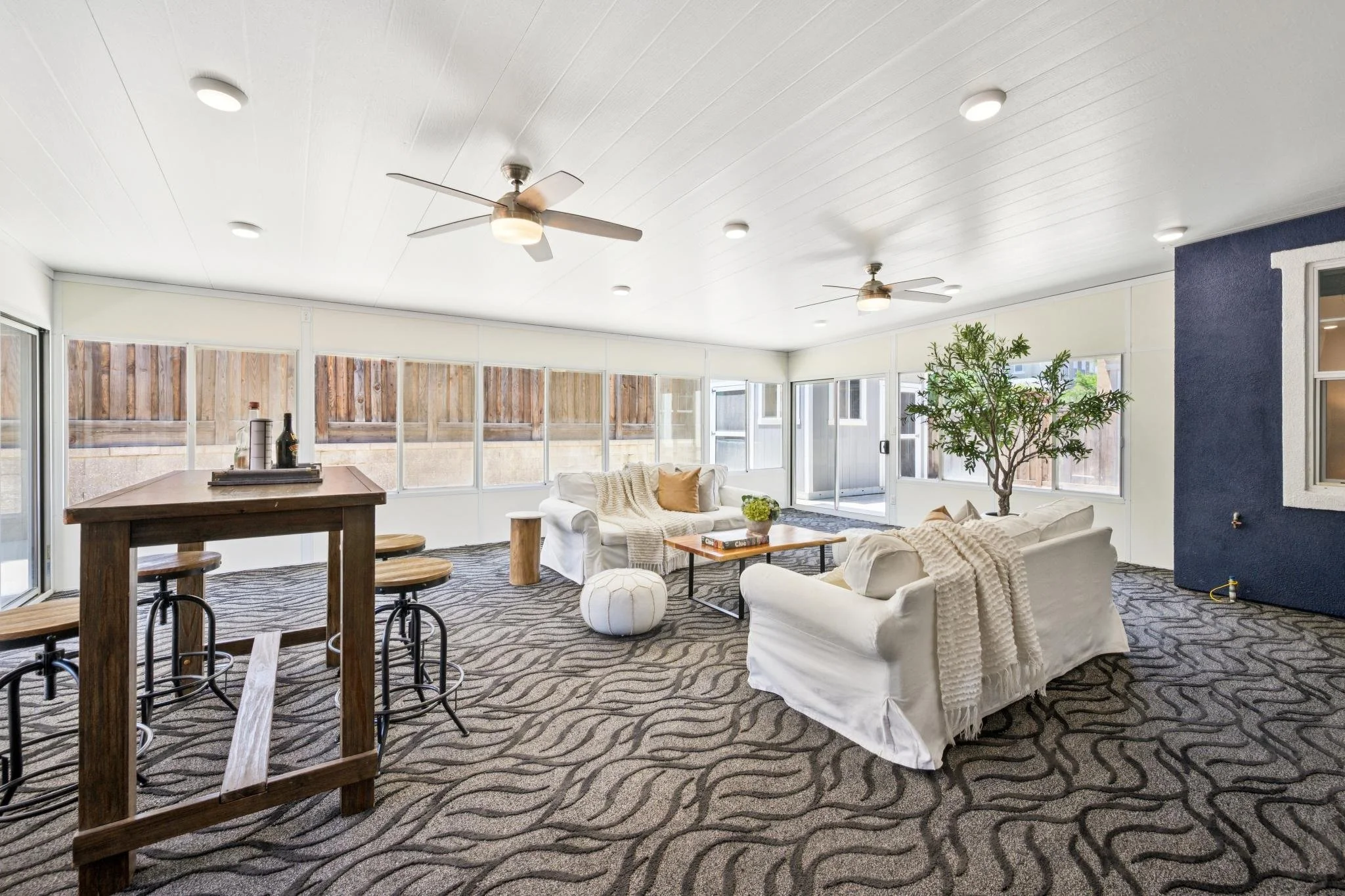 A bright sunroom with white walls and ceiling, ceiling fans, and large windows. There are white couches with pillows, a wooden table, and a potted plant. The floor has a dark patterned carpet.