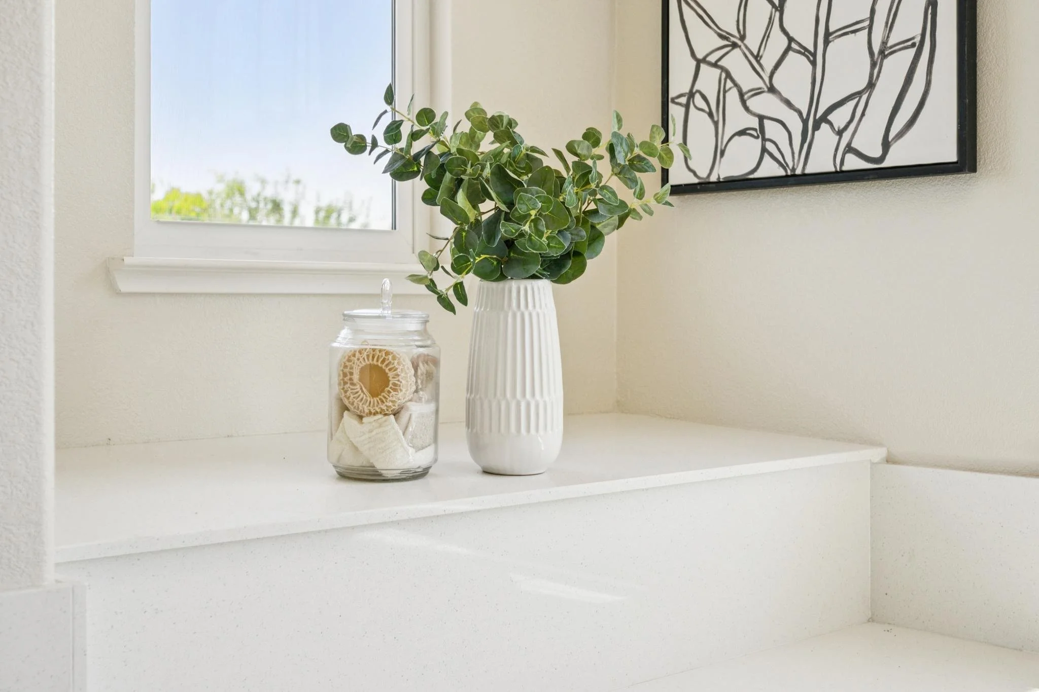 A windowsill with a potted plant, a jar with bath items, and a black and white floral line art wall hanging in a bright room.