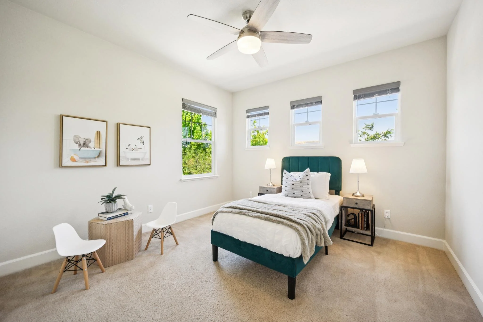 Bright bedroom with three windows, a teal upholstered bed, two bedside lamps, a nightstand, and wall art.