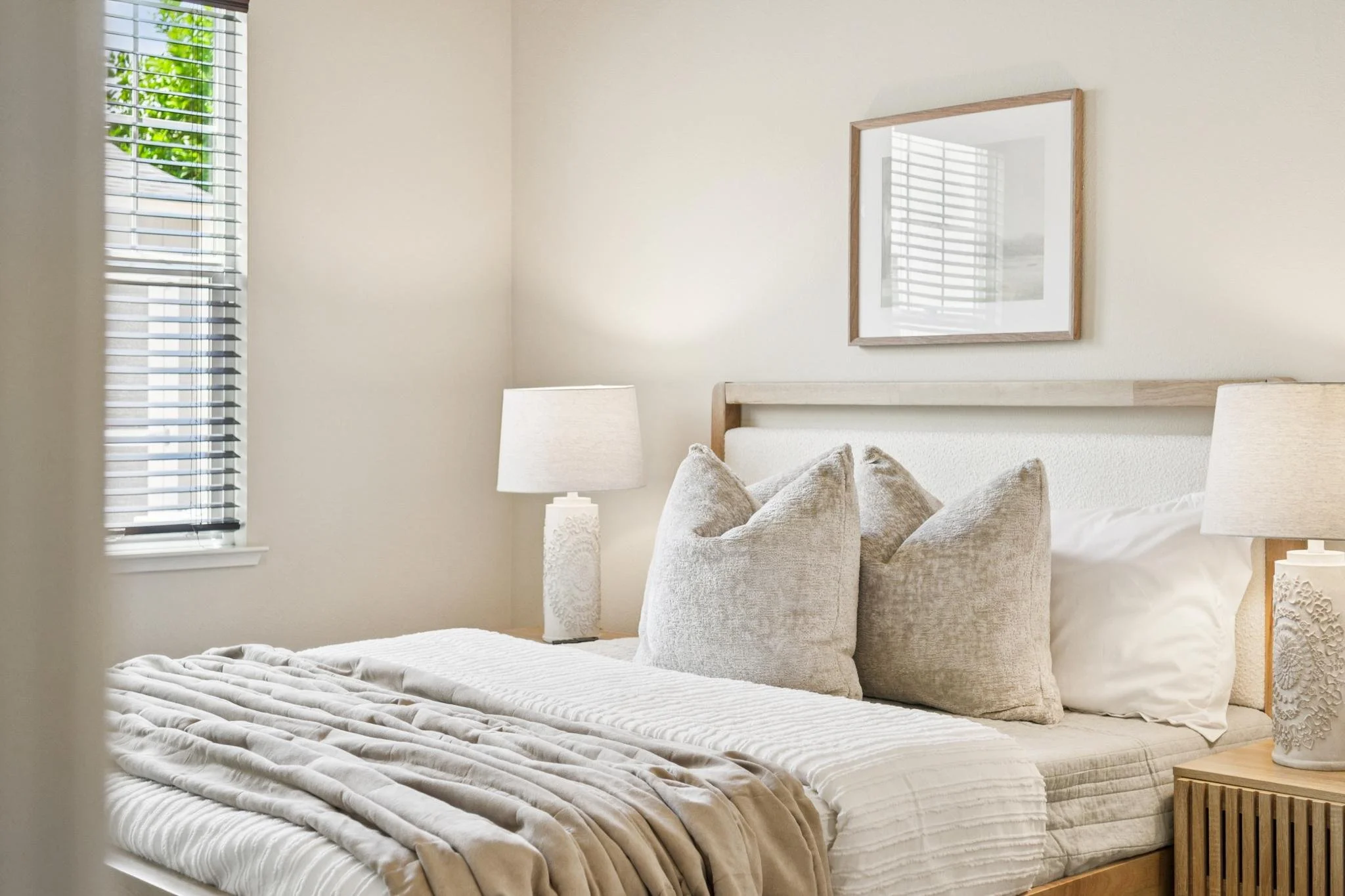 A neatly made bed with beige and white striped bedding, beige pillows, a wooden nightstand with a decorative white lamp, and a framed mirror hung on a light beige wall in a bright bedroom with a window.