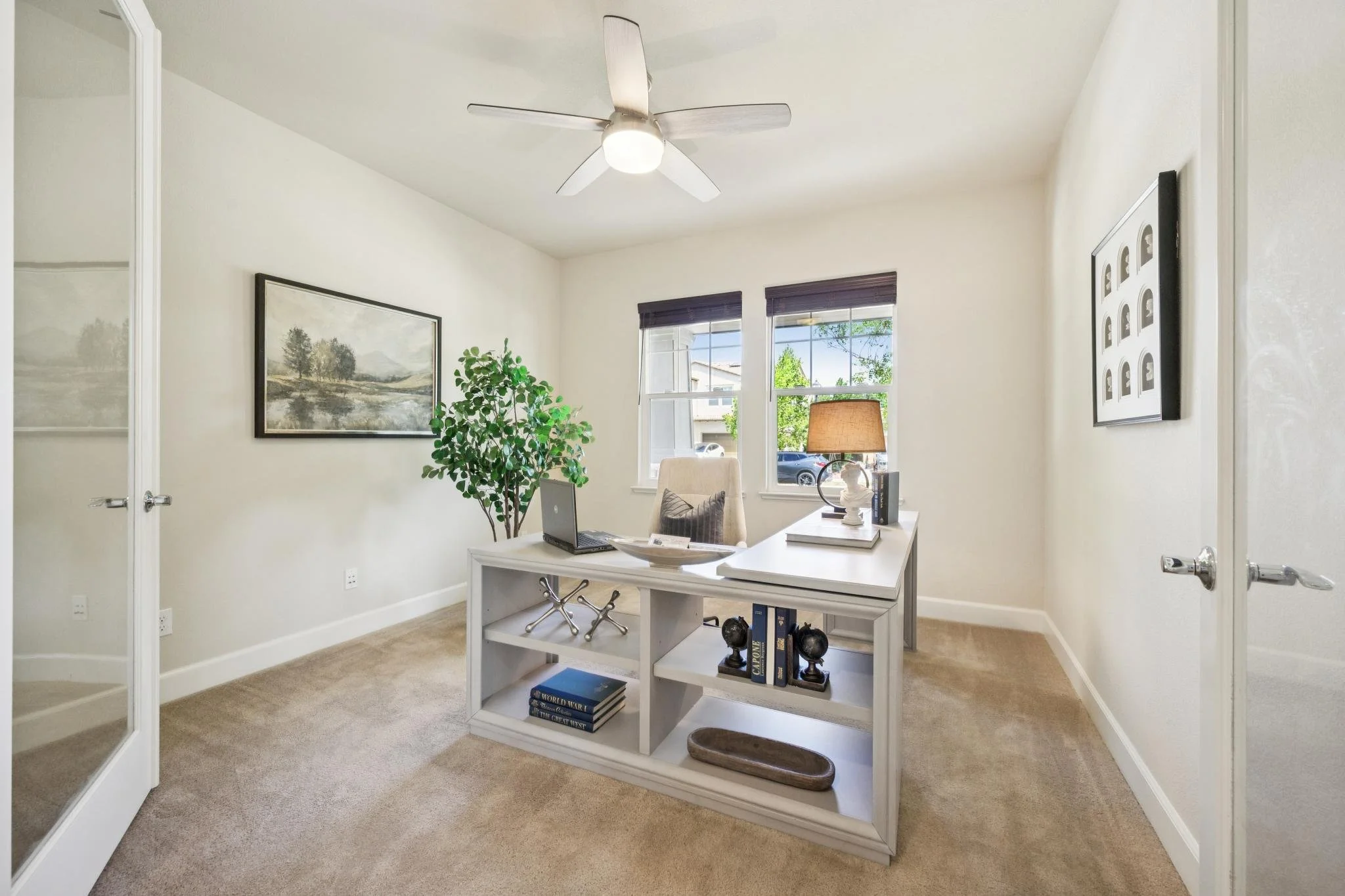 A bright office with cream-colored walls, beige carpet, and two windows with dark blinds letting in natural light. There is a ceiling fan with lights, a white desk with a laptop, decorative items, and books, a beige chair, a large framed landscape pa