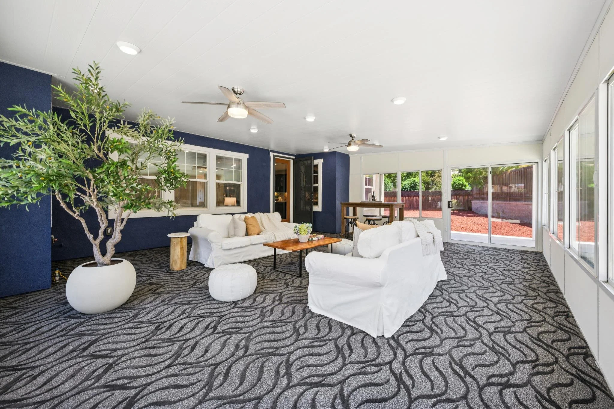 Living room with white furniture, a potted tree, large windows, and ceiling fans.