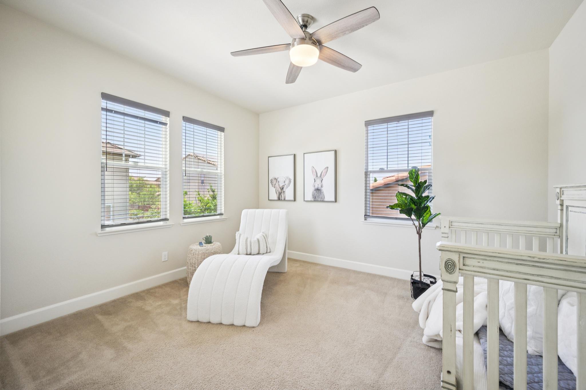 A bright, minimalistic living room with white walls, three large windows with blinds, a ceiling fan with wooden blades and a light fixture, a white upholstered lounge chair with a striped pillow, decorative animal art prints, and a green indoor plant