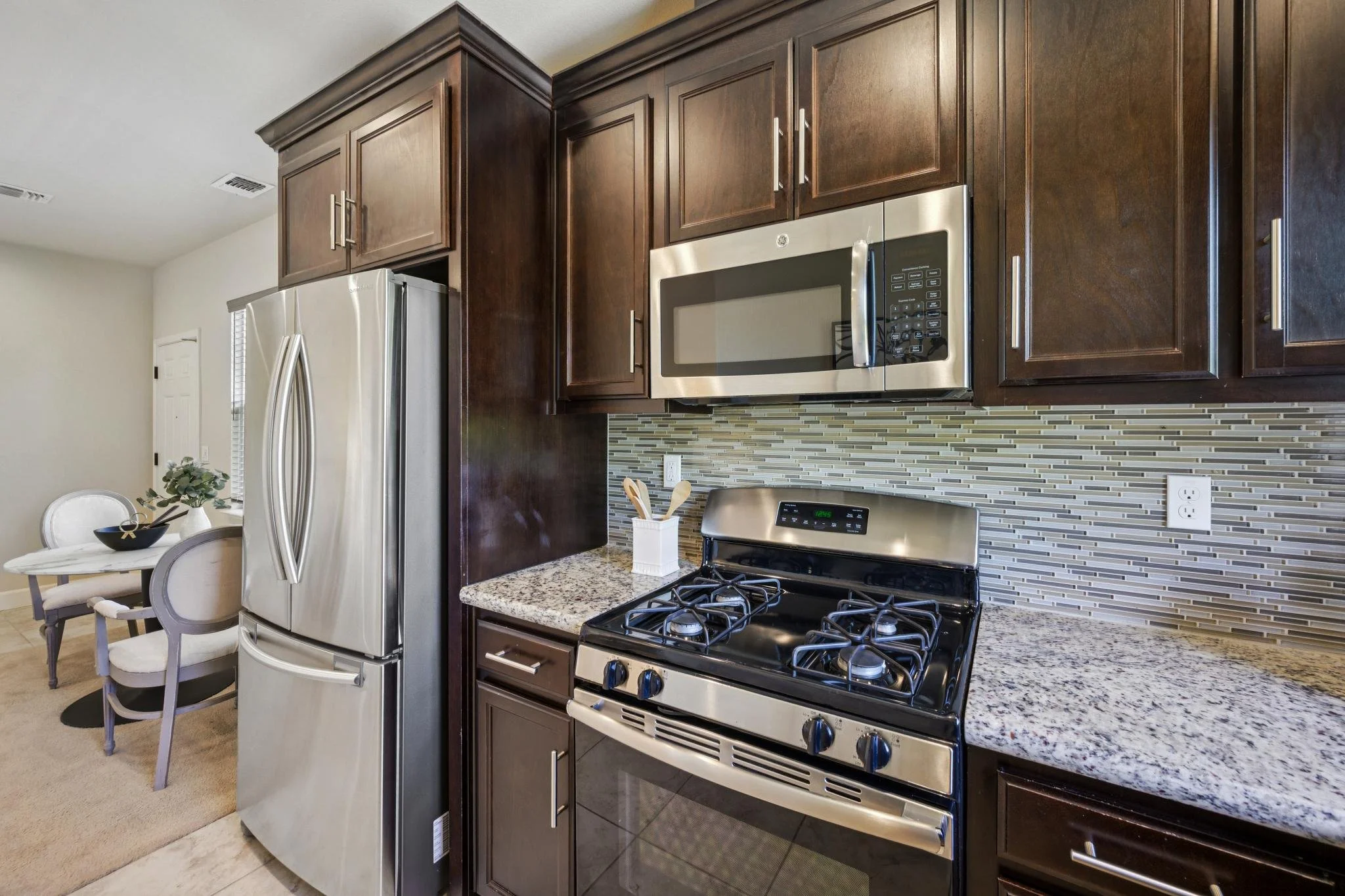 Kitchen with dark wood cabinets, stainless steel microwave and refrigerator, granite countertop, and a gas stove. Part of a dining area with a table and chairs is visible in the background.