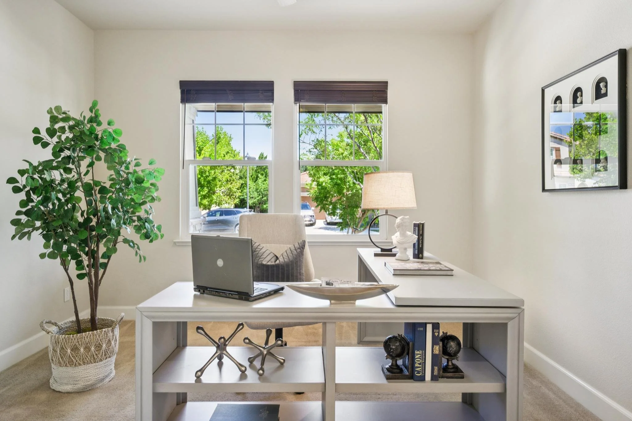 Home office with white desk, laptop, decorative objects, large potted green plant, window with blue blinds, framed artwork, and a lamp