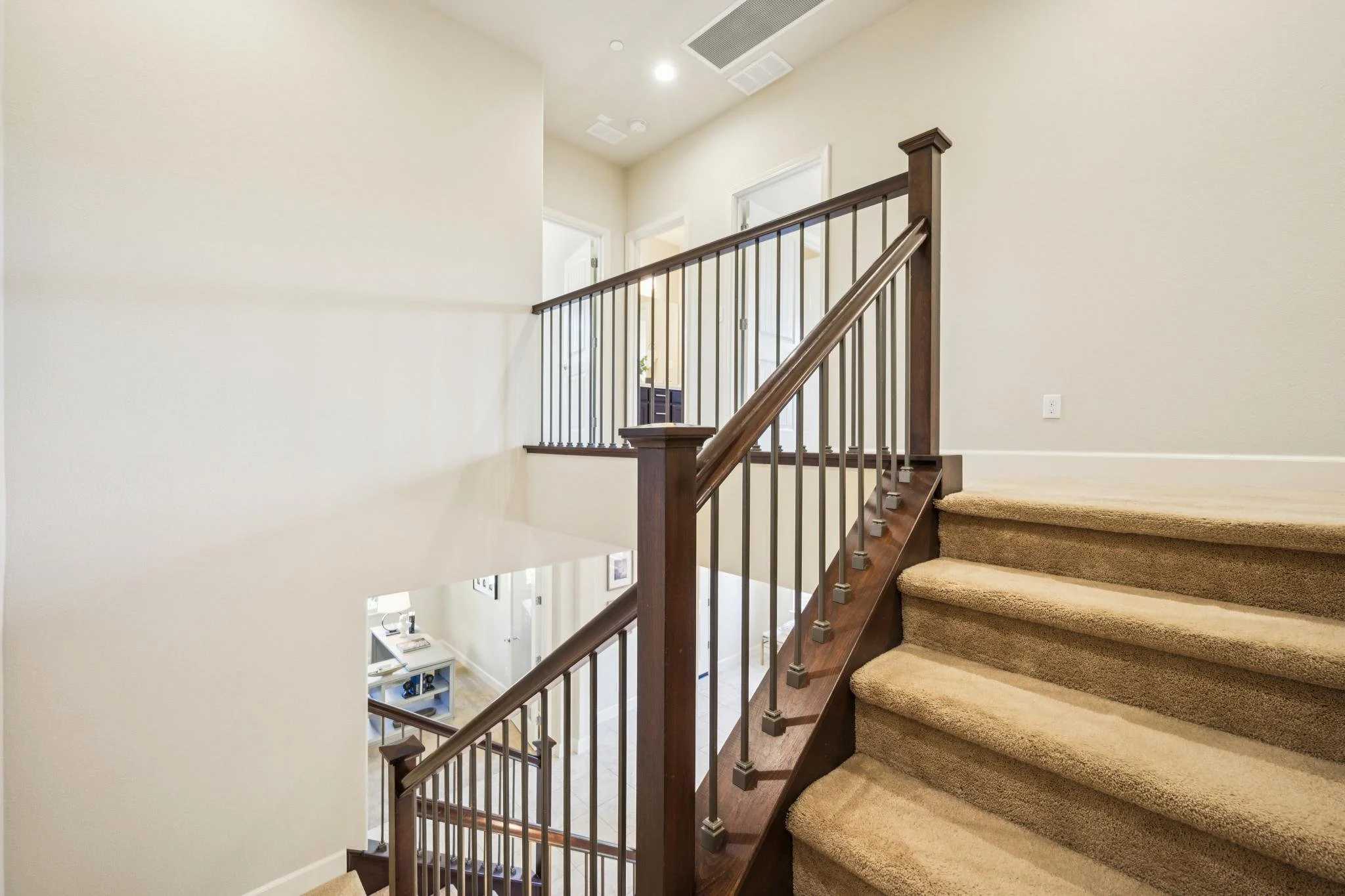 Interior view of a staircase with beige carpeted stairs, wooden handrail, and white walls, leading to an upper floor with multiple doorways visible.