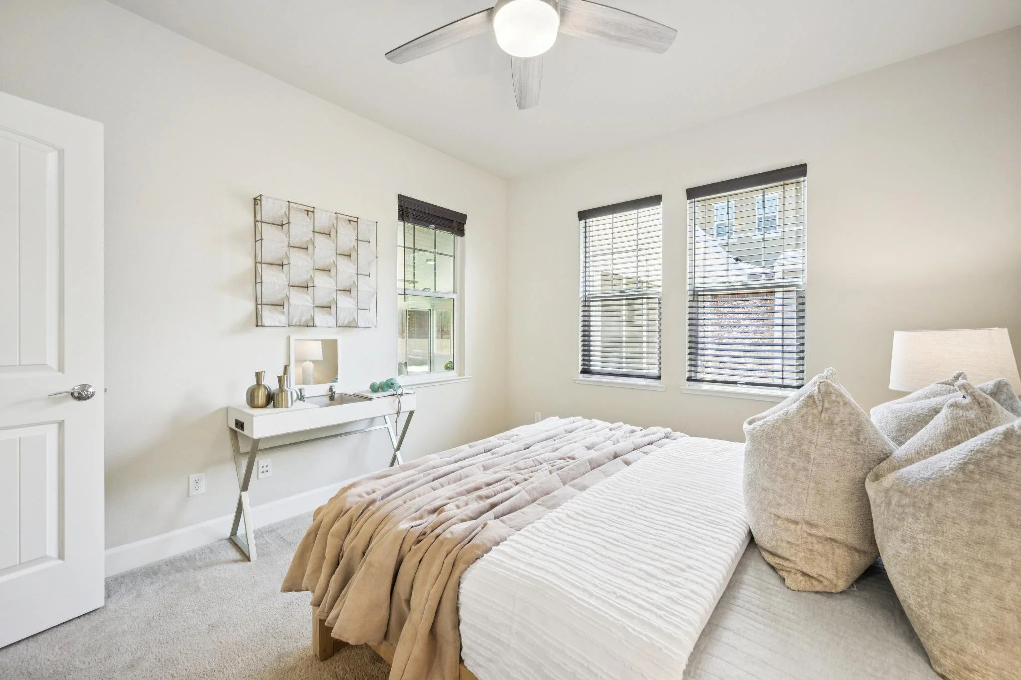 Bright bedroom with white walls, three large windows with black blinds, a ceiling fan with a light, a bed with beige and white bedding, and a white dresser with decorative vases.