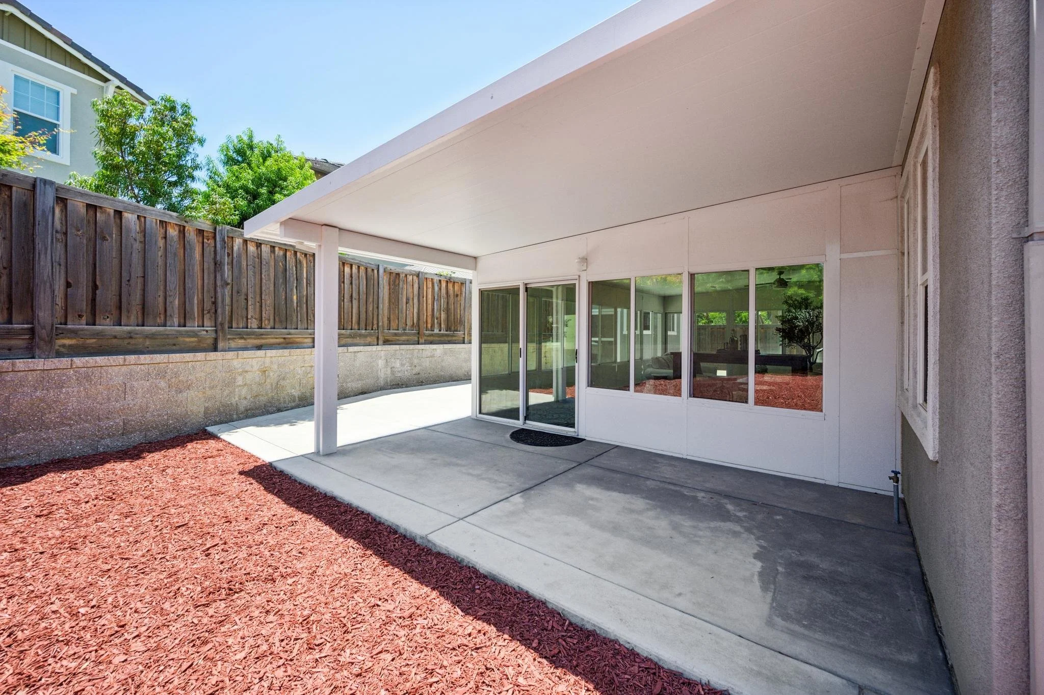Backyard patio area with a concrete floor, enclosed by a wooden fence and a glass door leading to the interior of a house. Red mulch surrounds the patio base. The house has white siding and large windows, with trees visible beyond the fence. The pati