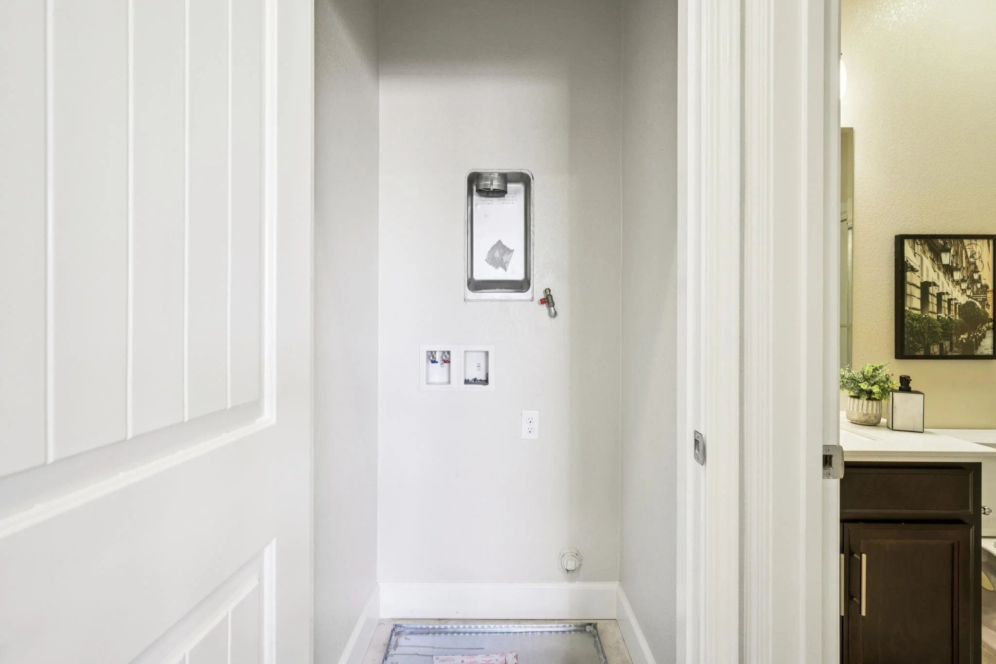Empty laundry room with a drain in the floor, wall outlets, and an elevated water connection with a water valve, adjacent to a kitchen or dining room with a framed picture on the wall, a potted plant, and a small decorative item.