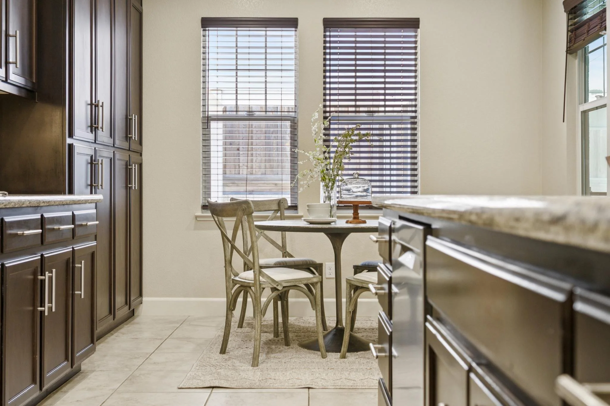 Kitchen area with dark wooden cabinets, a small round dining table with two chairs, a vase with white flowers, windows with blinds, and tiled flooring.