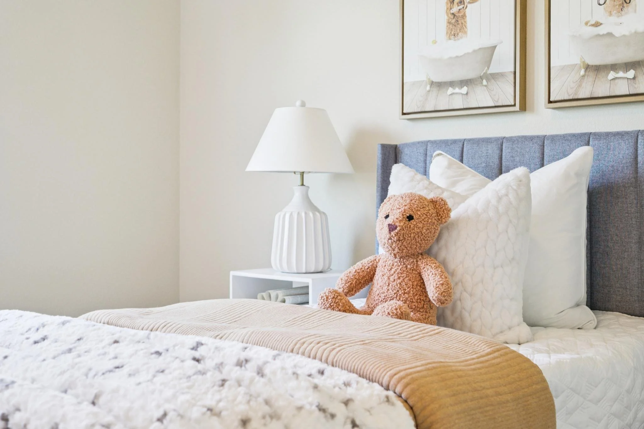 A cozy bedroom corner featuring a plush teddy bear sitting on a bed with white and beige bedding, a white bedside table with a lamp, and framed artwork hanging on the light-colored wall.