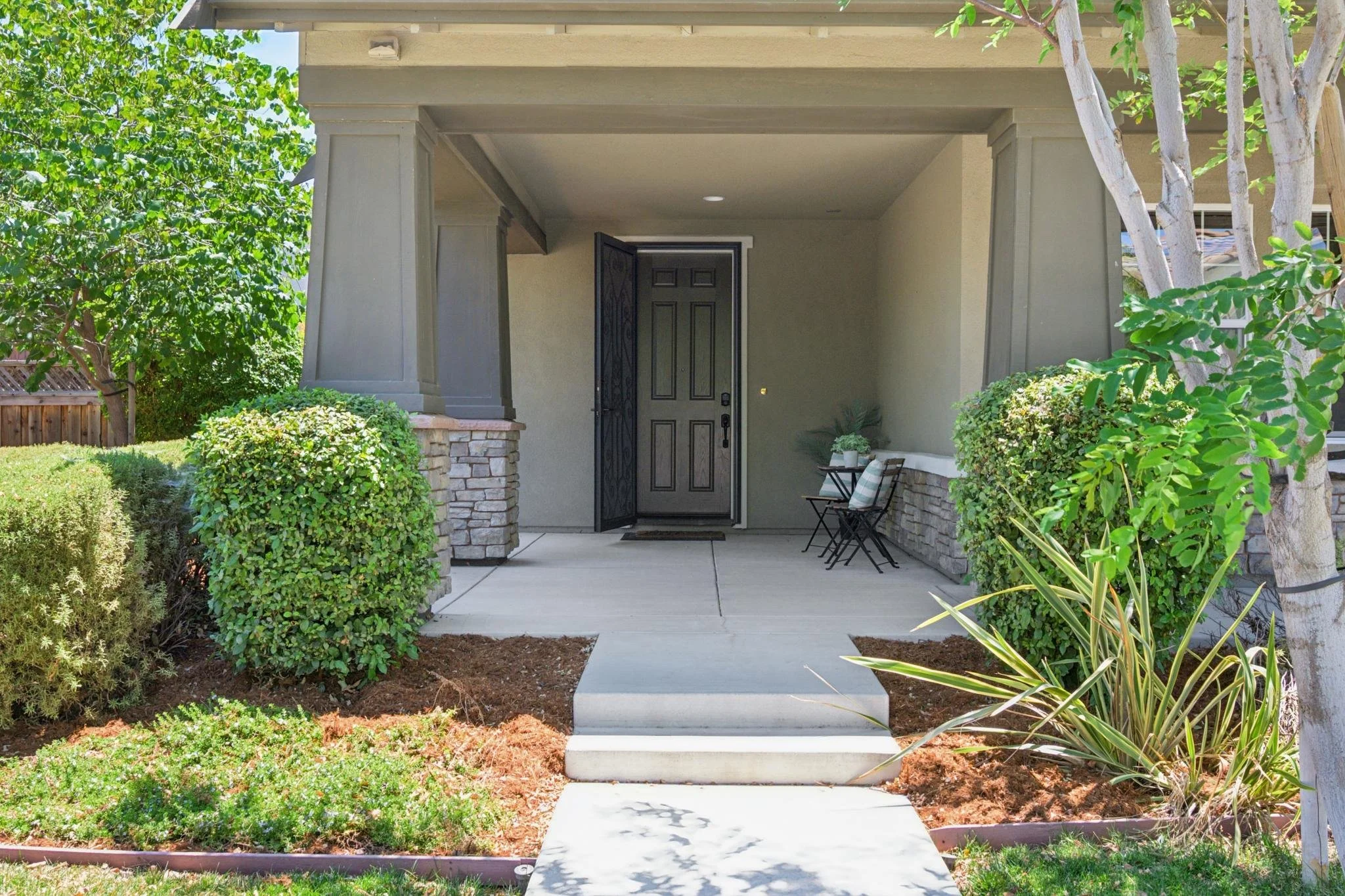 Front porch of a house with a black door, two chairs, surrounded by green bushes and small trees, with a sidewalk leading to the door.