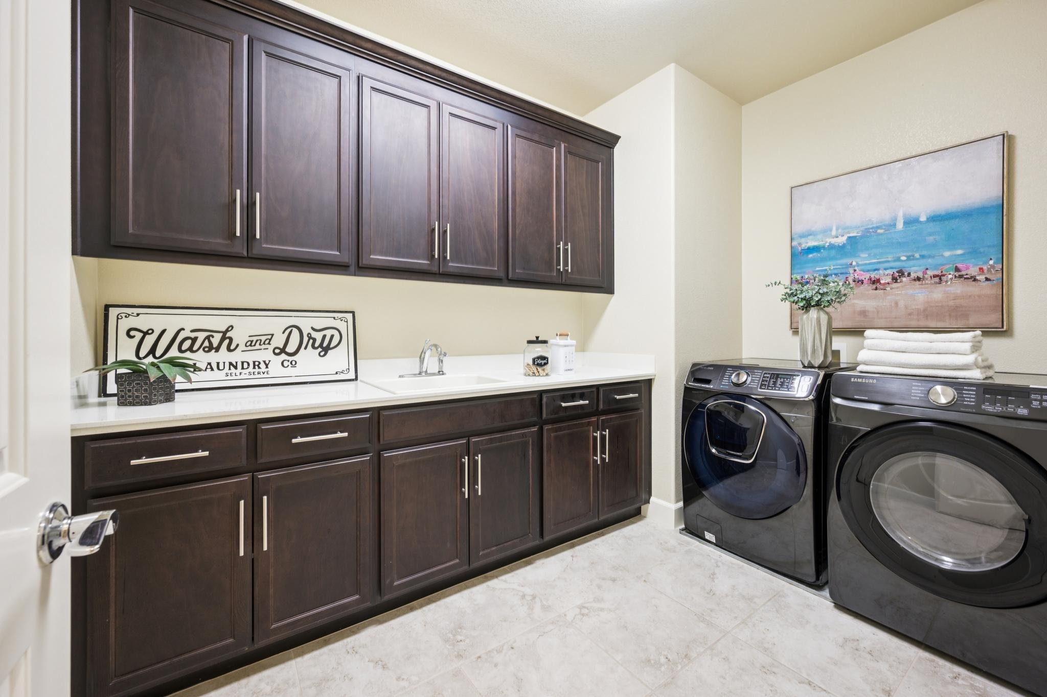 A laundry room with dark wood cabinets, a white countertop, a sink, and laundry supplies. There are two front-loading washers and a dryer, a vase with flowers, a stack of folded towels, and a large beach-themed painting on the wall.