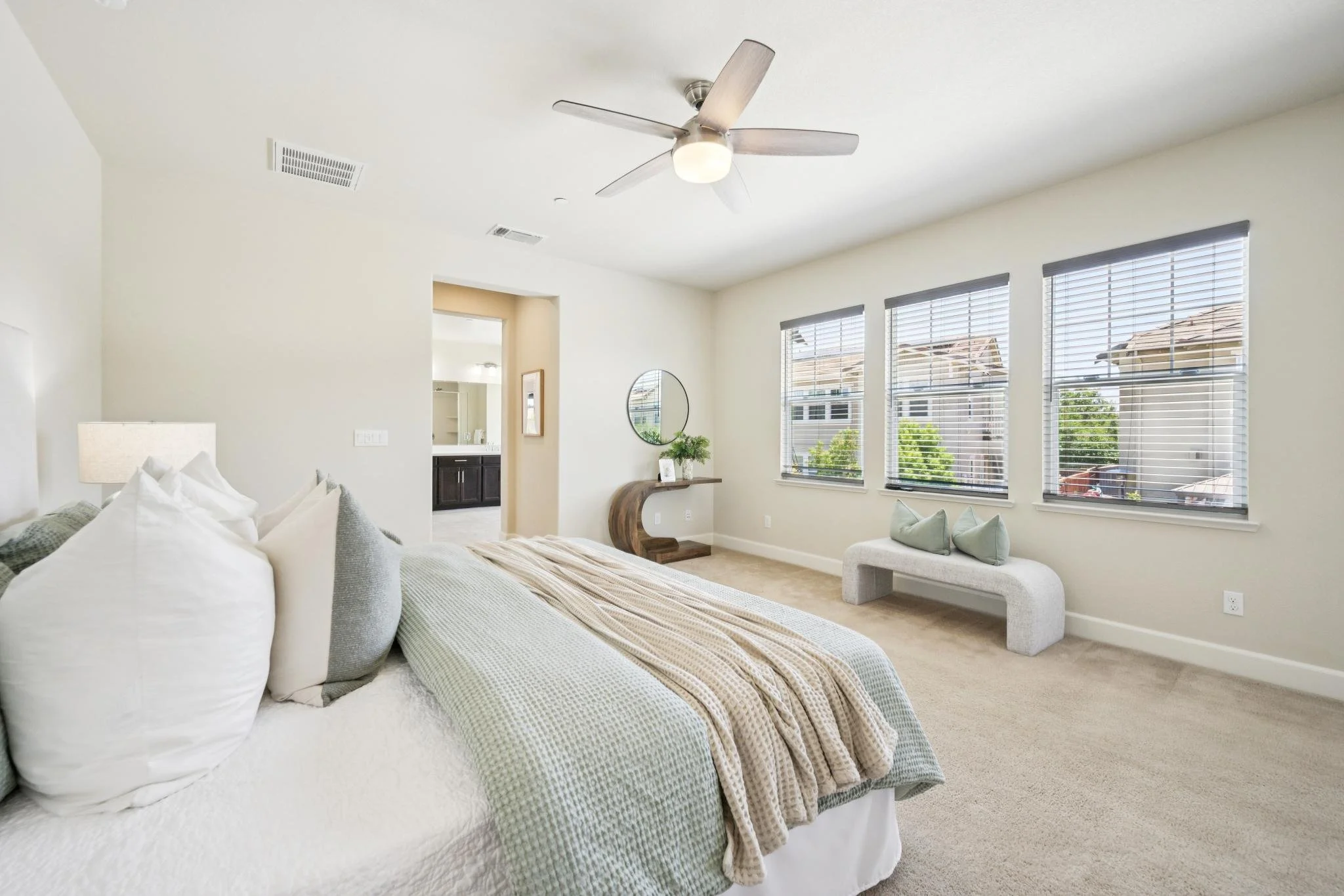 Bright bedroom with large windows, a bed with white linens, gray and beige throw pillows, a beige blanket, a small side table with a white lamp, a mirror, a wooden console table with a plant, and a ceiling fan.