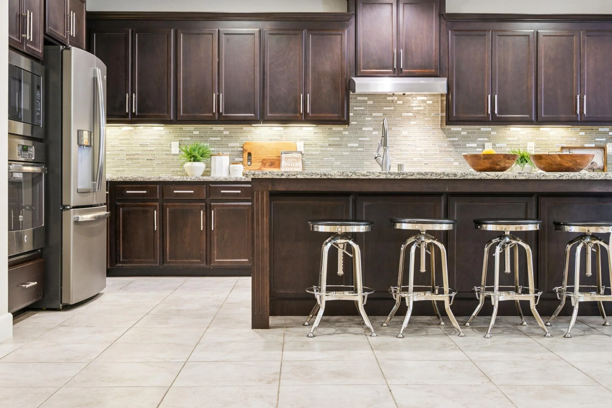 Modern kitchen with dark wooden cabinets, stainless steel appliances, a tiled backsplash, and a granite countertop. There is a kitchen island with four chrome barstools, decorative bowls, and small plants.