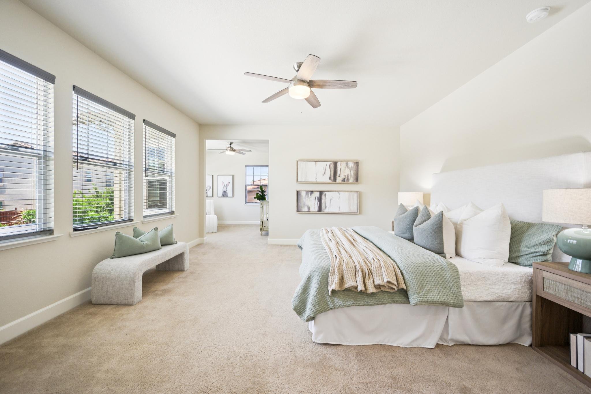 A spacious bedroom with a large bed, nightstand, and window with blinds. Modern decor with framed artwork and a footstool near the window. Ceiling fans are visible in the adjoining rooms.