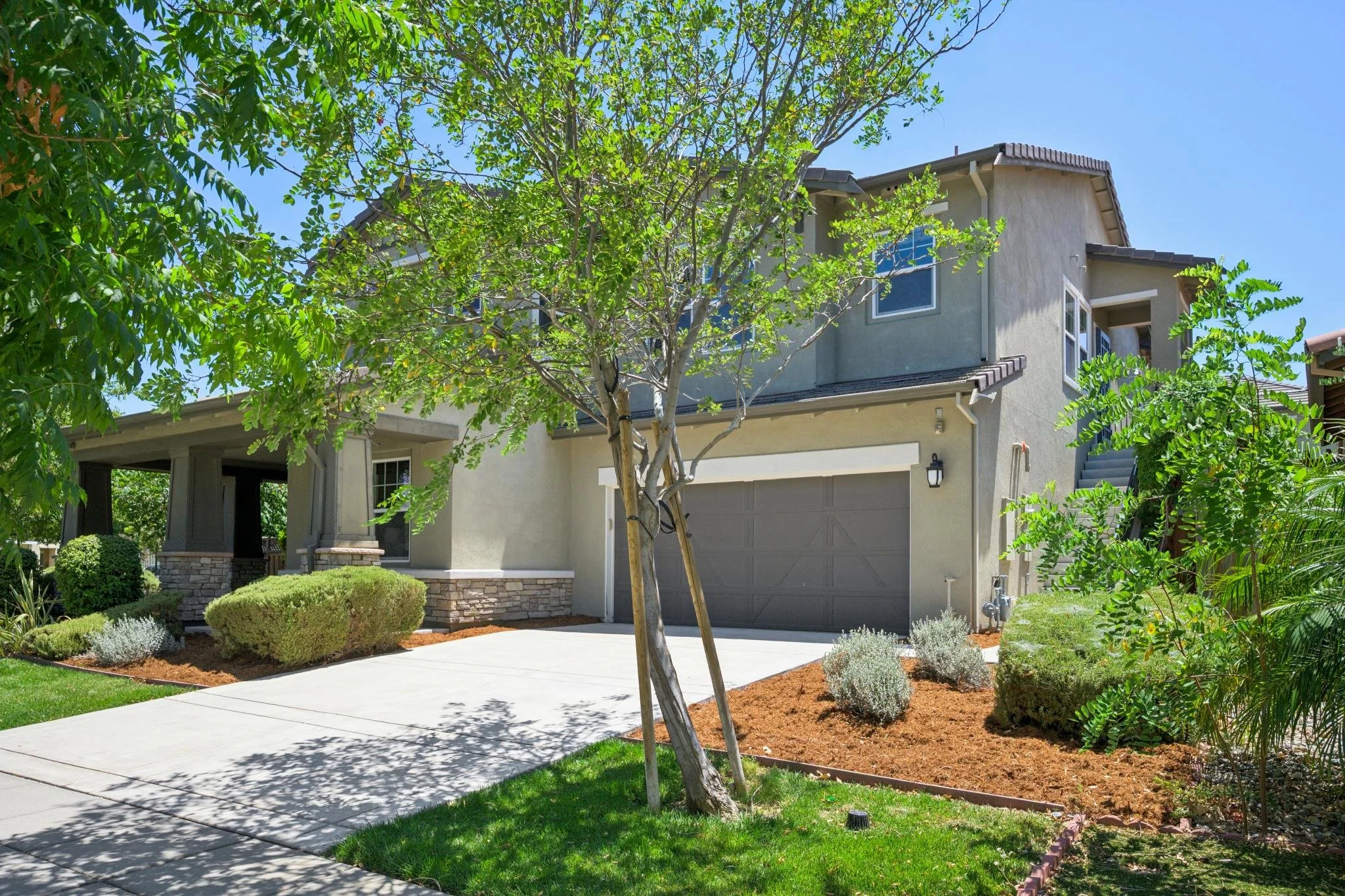Front yard of a house with trees, shrubs, and a concrete walkway leading to the garage. The house has light-colored walls and multiple windows. The landscaping is well-maintained with patches of grass and mulched garden beds.