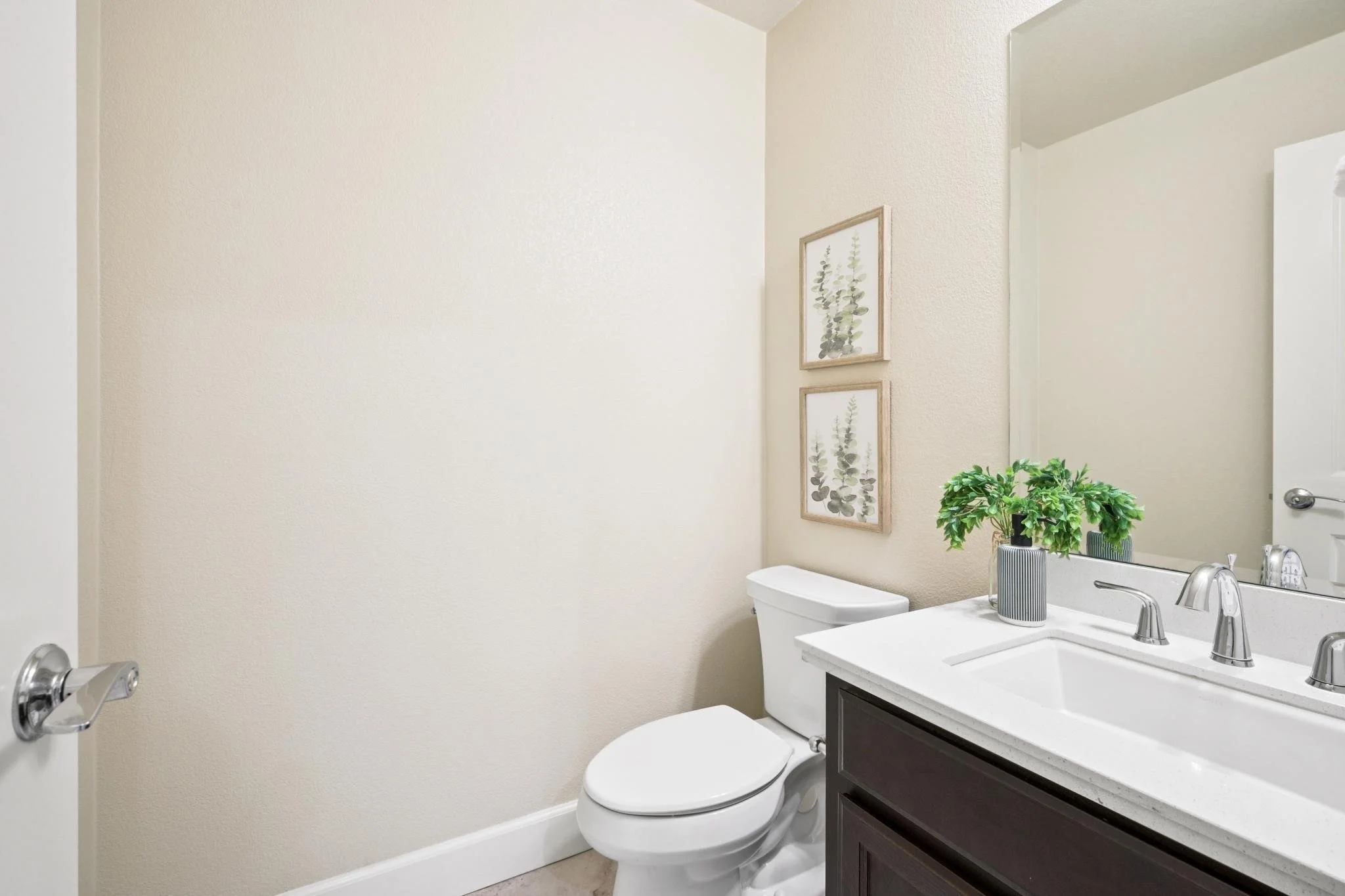 A small bathroom with a white toilet, a white sink with a stainless steel faucet, a dark wooden vanity, and a mirror above the sink. There are two framed botanical prints on the beige textured wall and a potted plant on the sink counter. The bathroom