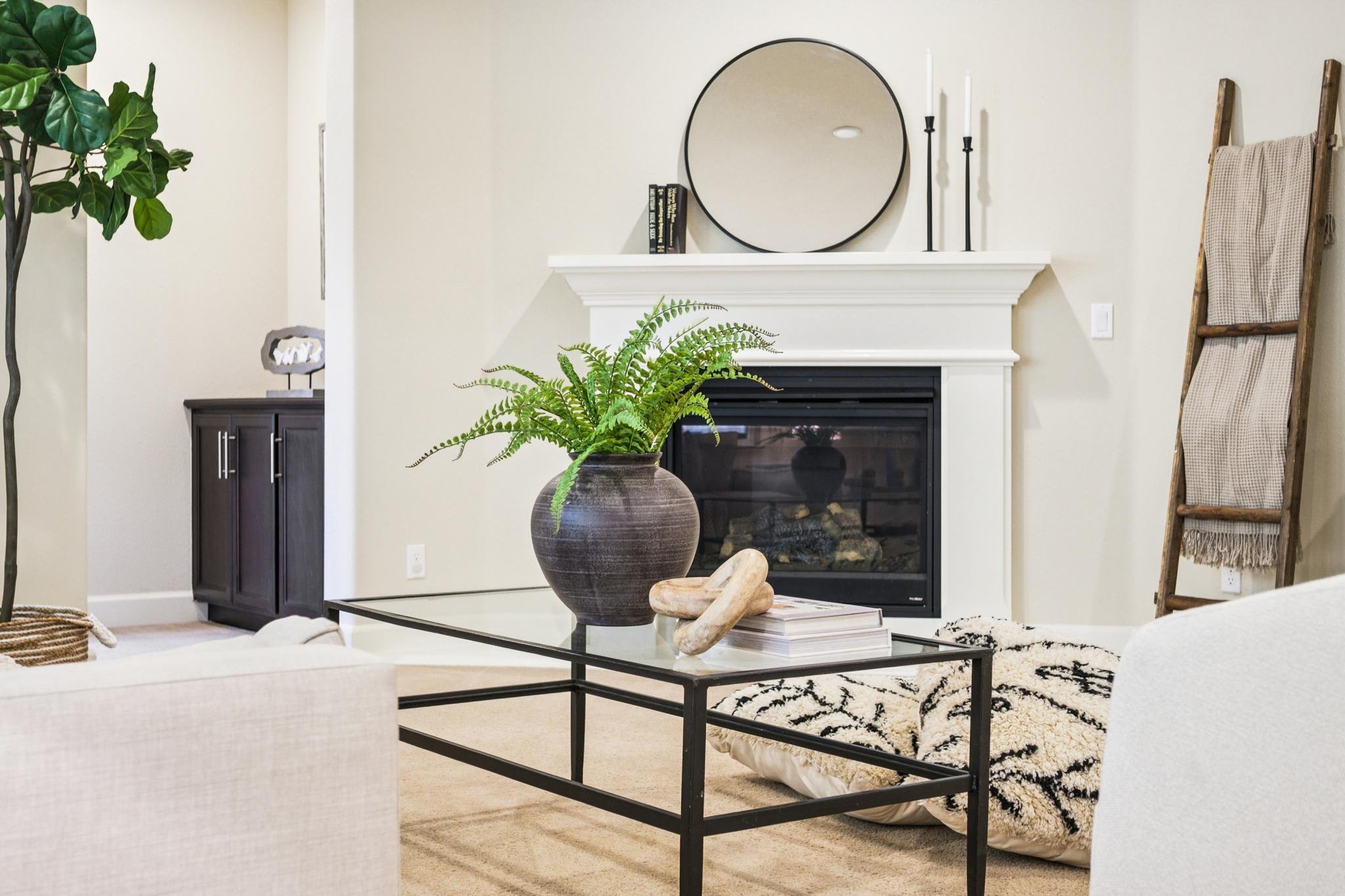 Living room with a white fireplace, a round mirror above it, black candlesticks, and decorative books. There is a potted fern in a dark brown vase on a glass-top coffee table, with a white and black patterned rug and a beige sofa.