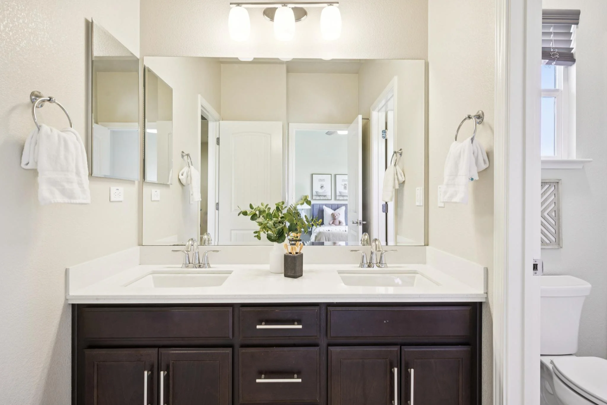 Bathroom vanity with dual sinks, dark wood cabinet, large mirror, decorative plant, and modern light fixture.
