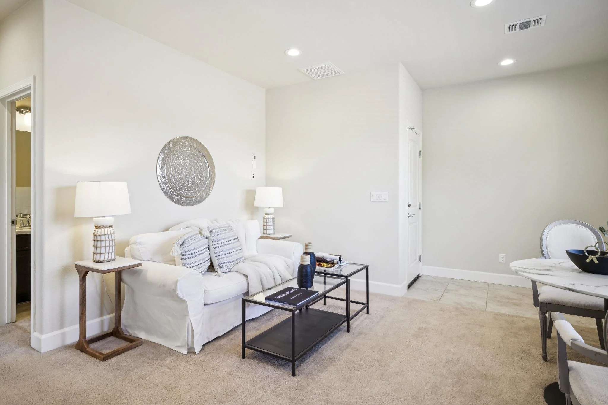 Living room with white walls, beige carpet, a white couch with decorative pillows, a wooden side table with lamps, a black coffee table, and decorative vases, with an open doorway leading to another room.