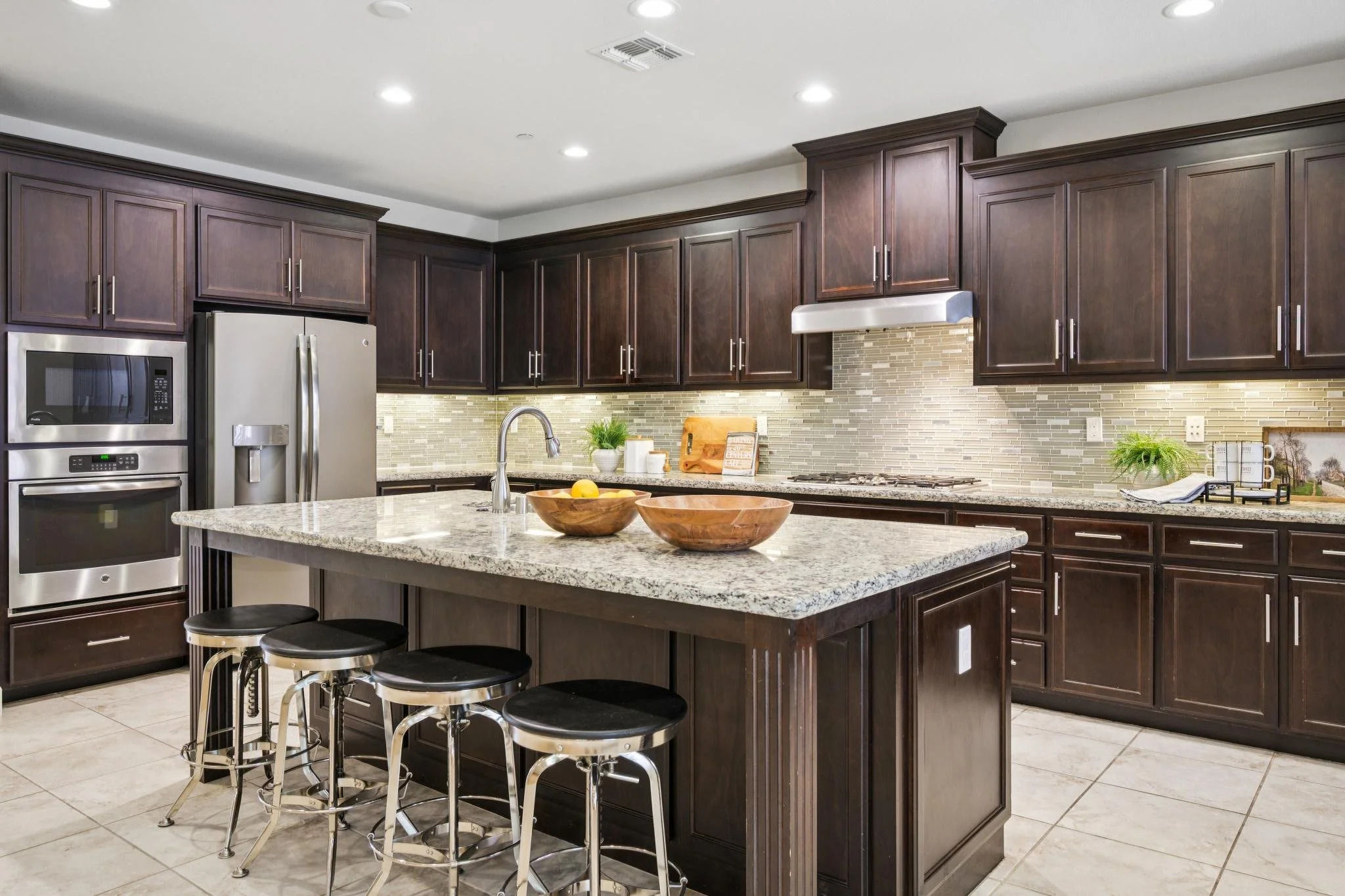 Modern kitchen with dark wooden cabinets, granite island, stainless steel appliances, beige tiled floor, and decorative backsplash.