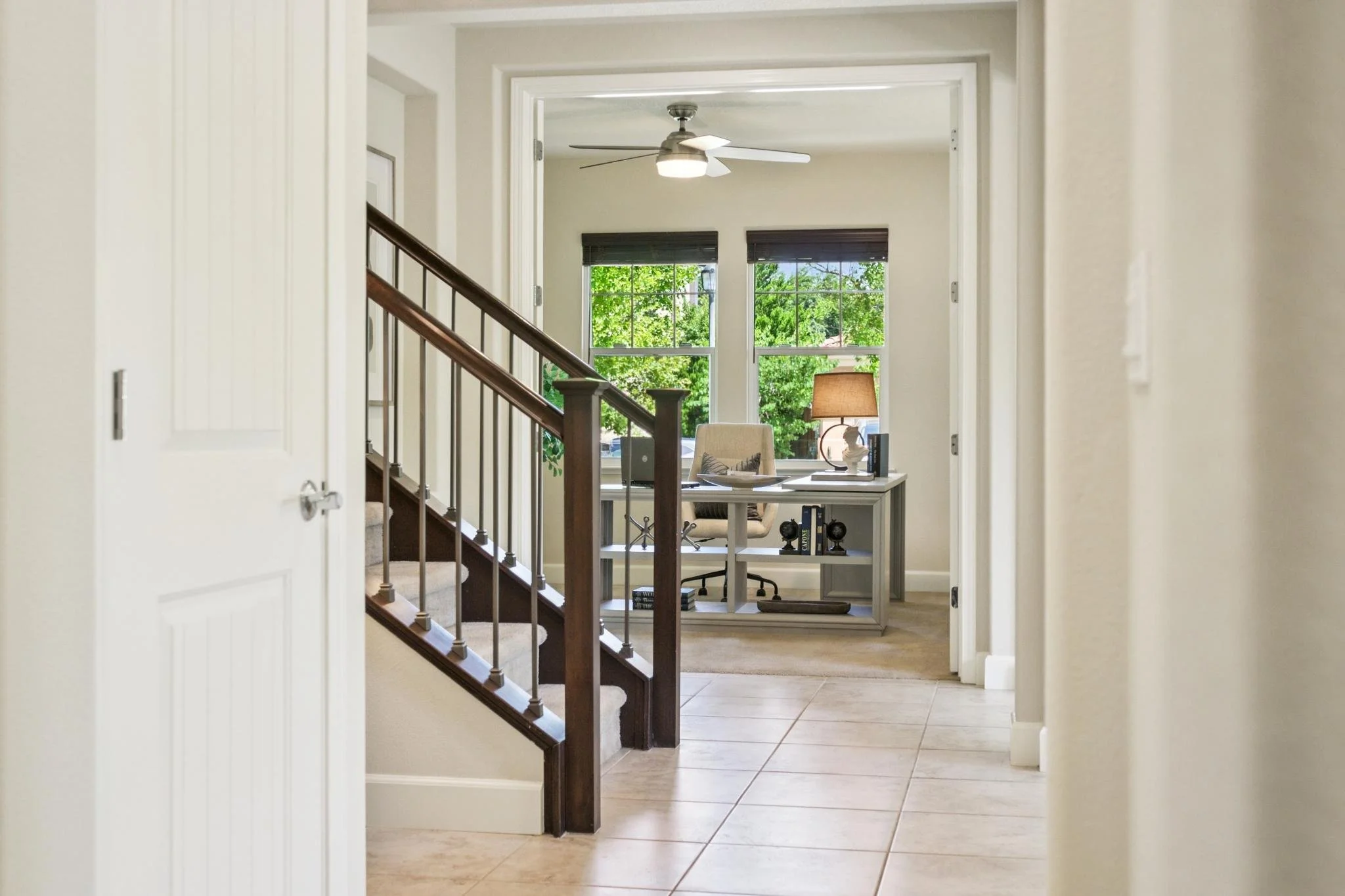 View from hallway into home office with large windows, ceiling fan, beige walls, white desk, beige chair, table lamp, and decorative items.