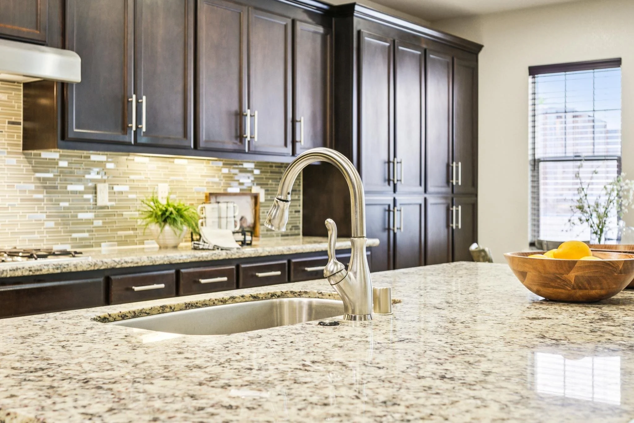 A kitchen with dark wood cabinets, a granite countertop, a modern faucet, a wooden bowl with fruit, and a window with blinds.