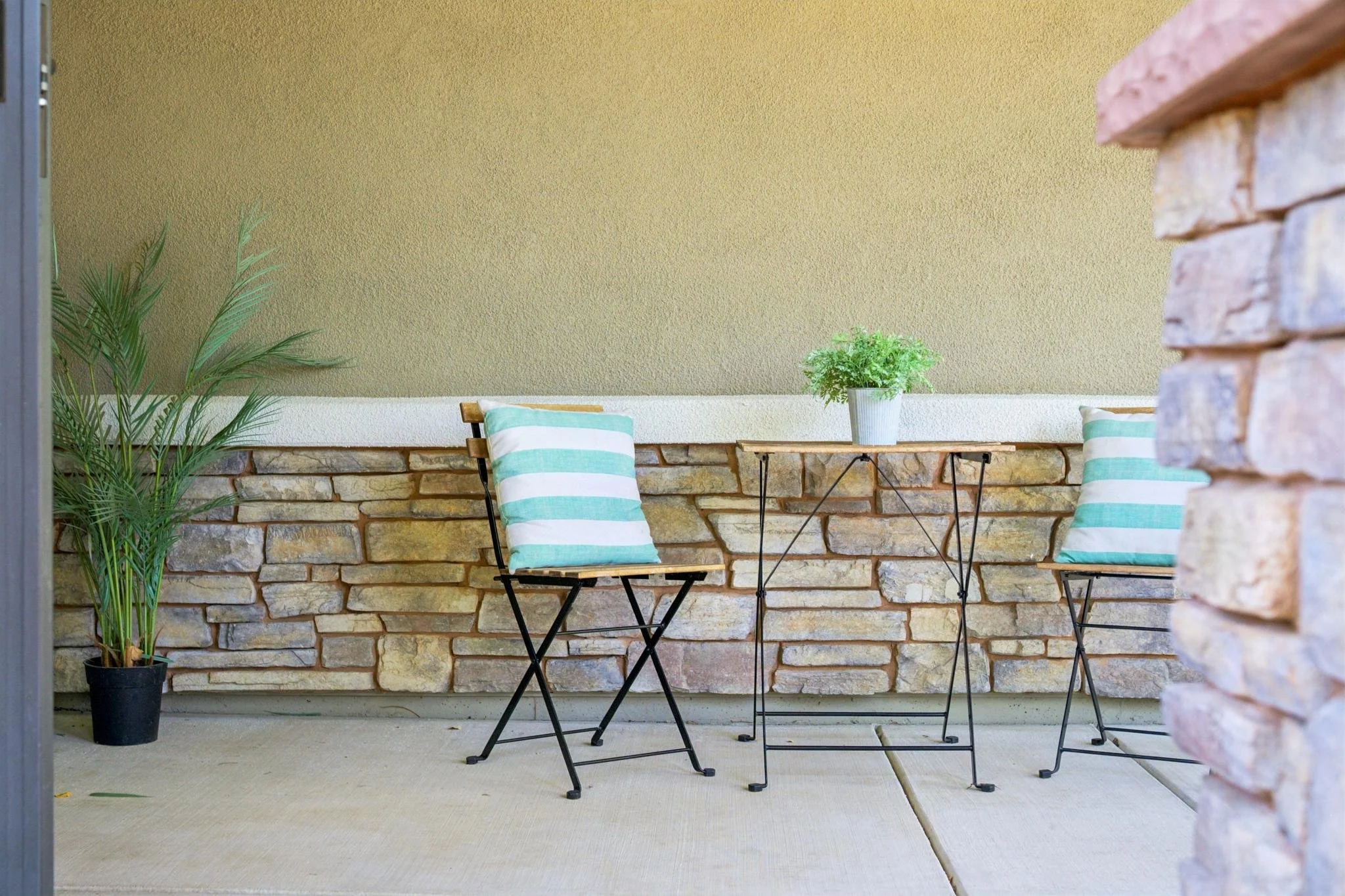Outdoor patio area with two black folding chairs, a small wooden table, and decorative pillows with teal and white stripes, against a stone wall with a beige and yellow textured wall in the background, and a potted green plant on the table.