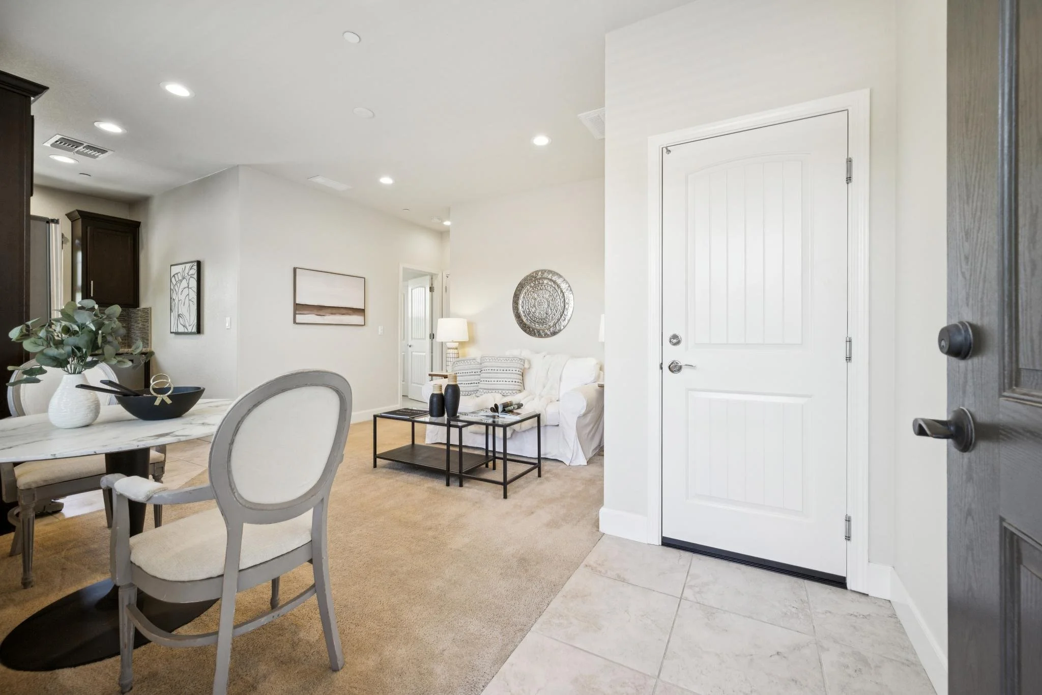 View of a living room and dining area with white walls, a white sofa, a side table with black decorative vases, and wall art, with a doorway partly open revealing tiled flooring near the entrance.