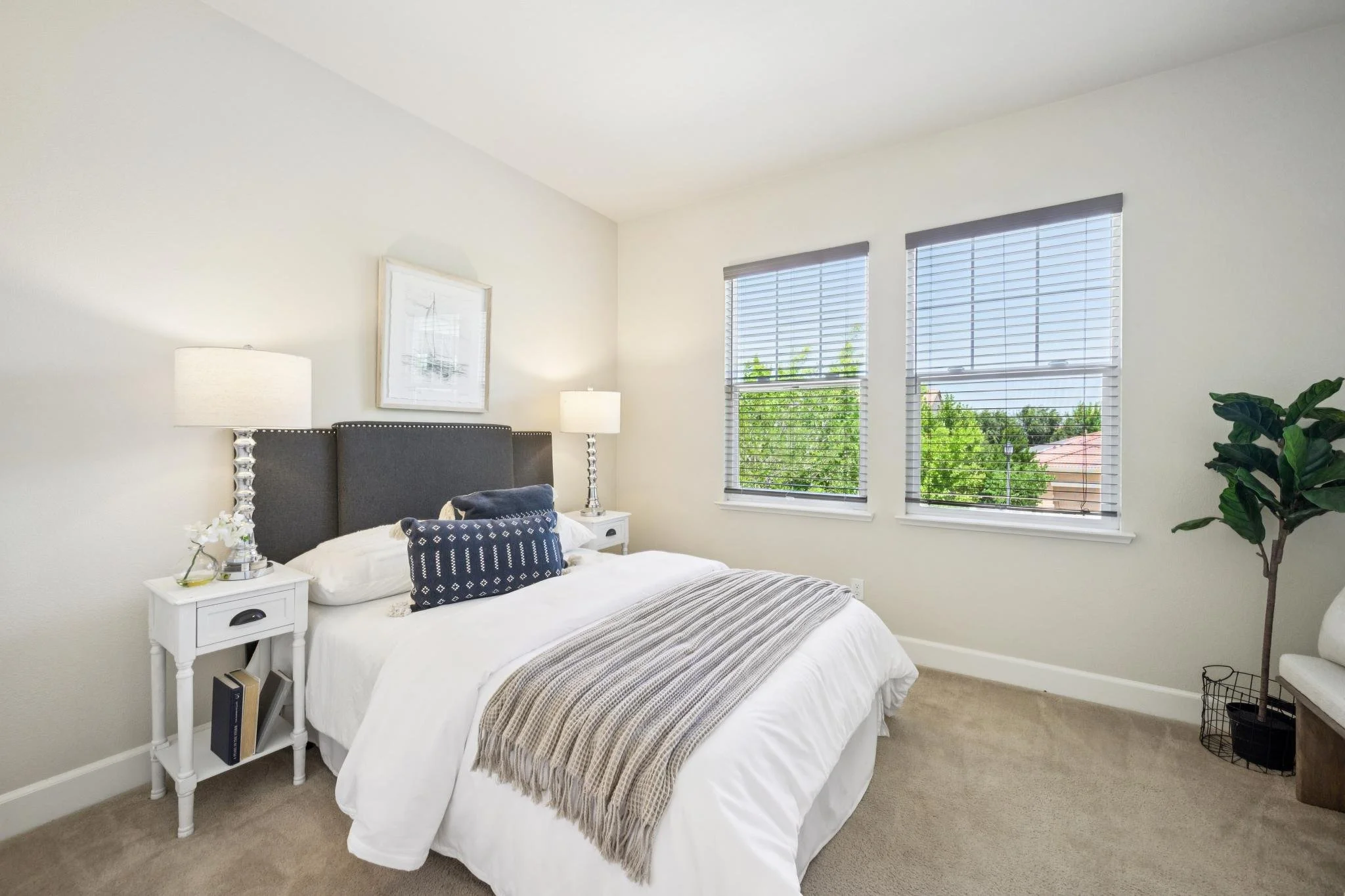 A bedroom with a bed, white bedding, navy blue decorative pillows, and a gray upholstered headboard. Two white nightstands with lamps are beside the bed. There are two large windows with white blinds and a view of trees outside. A framed picture hang
