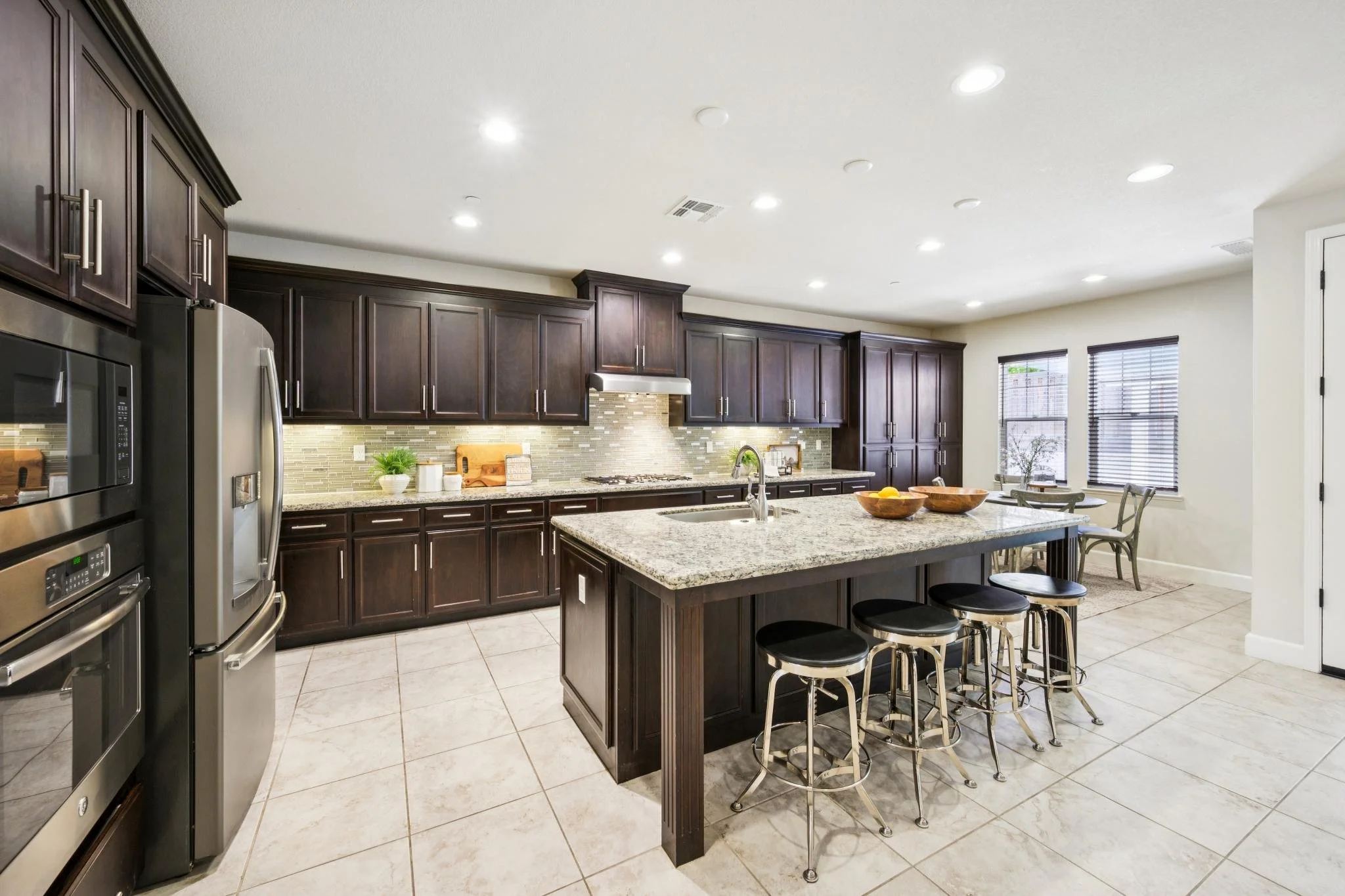 Modern kitchen with dark wooden cabinets, granite countertops, stainless steel appliances, and a breakfast bar with five stools. Bright lighting and windows with blinds let in natural light.