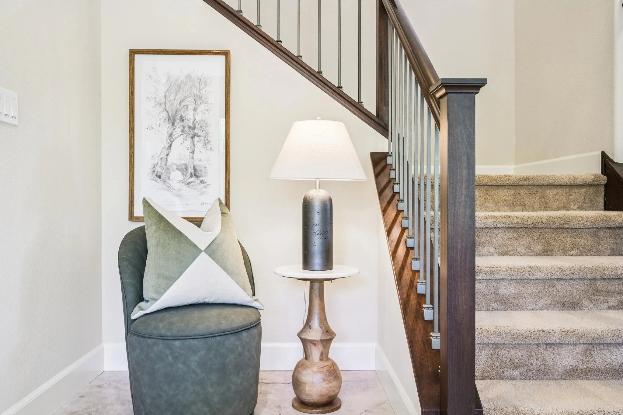 A close-up of a living room corner featuring a dark gray upholstered chair with a decorative pillow, a wooden side table with a beige ceramic table lamp, a large framed black-and-white tree artwork on the wall, and a staircase with beige carpeted ste