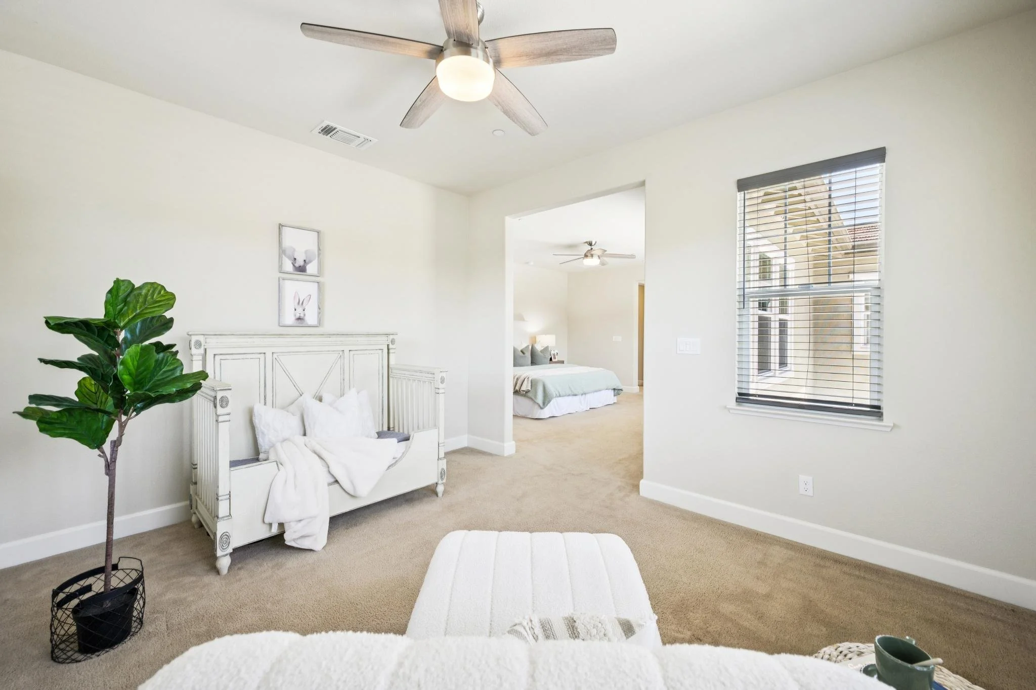 A bright, modern bedroom with white walls, large window with blinds, ceiling fans, and a bed in the adjoining room.